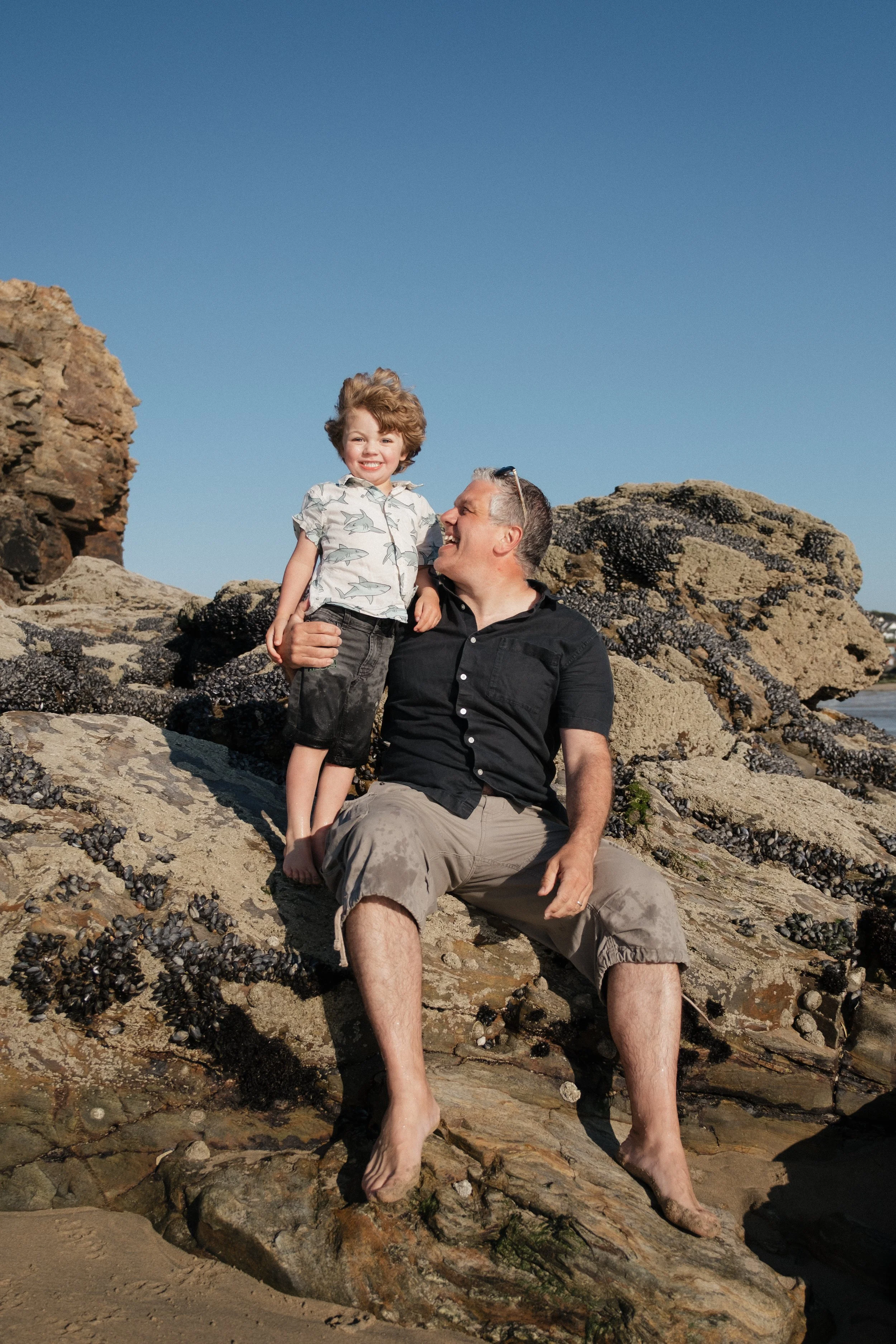 A man and a young boy sitting on rocks at the beach, smiling and enjoying a sunny day.