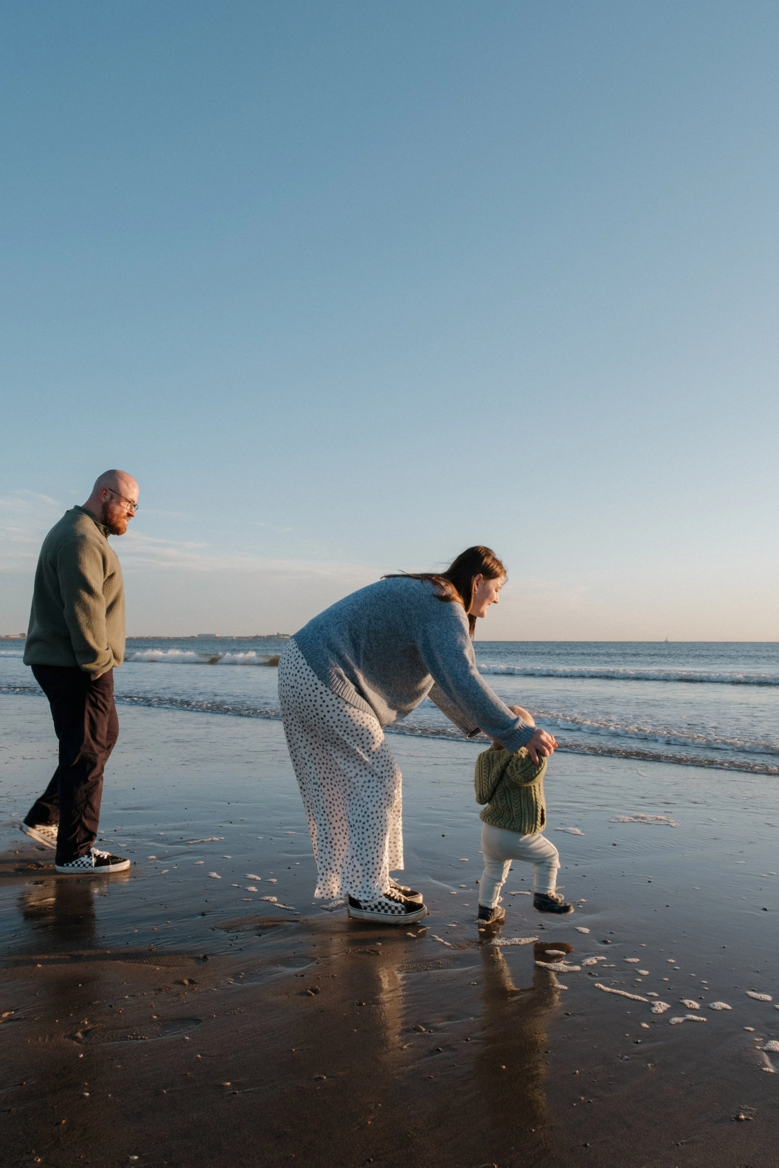 A family of three walking on the beach during sunrise, the mother is guiding a young child who is walking toward the water, and the father is walking behind them.