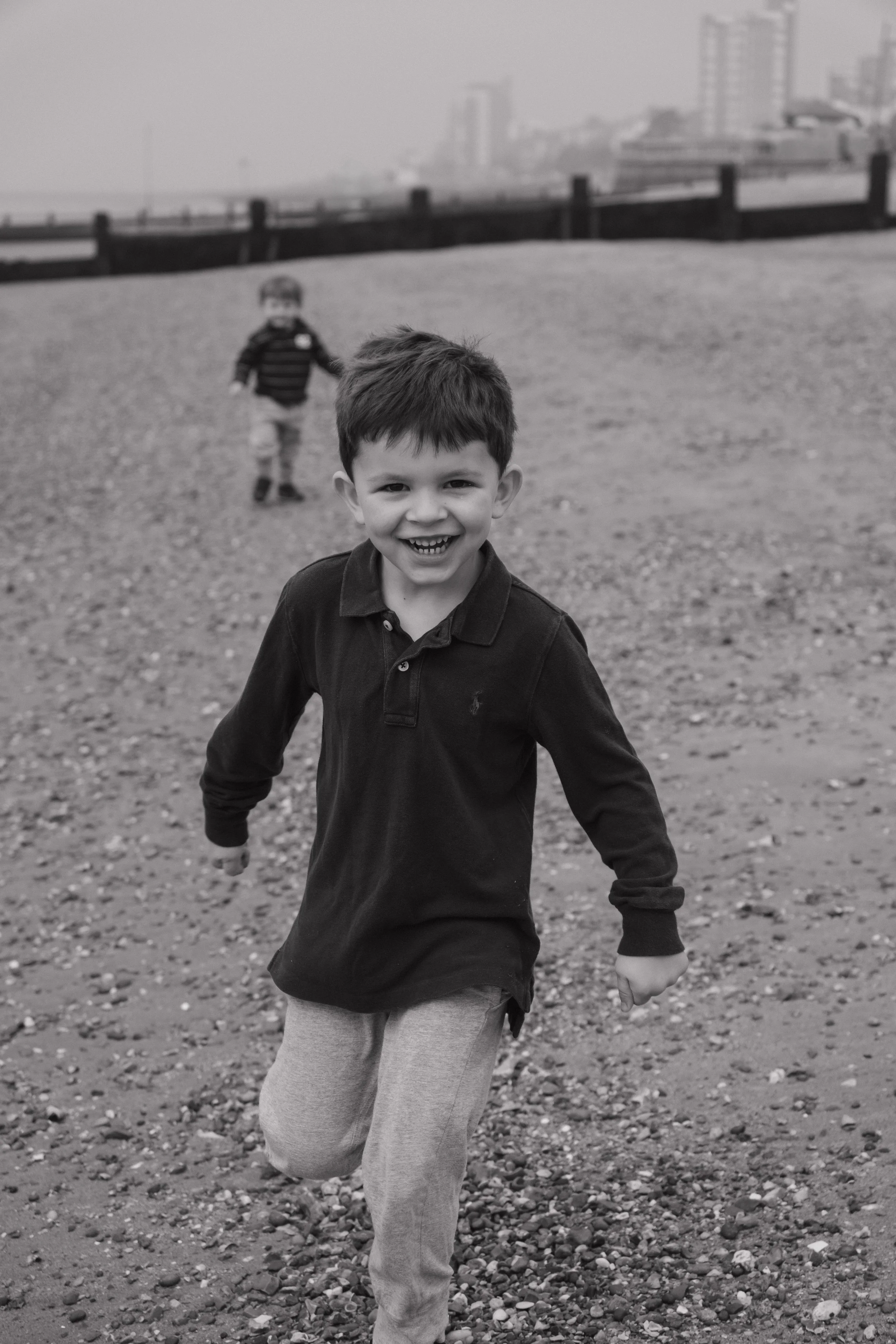 Young boy running on a gravel beach, smiling, with a younger child in the background, city skyline in the distance.