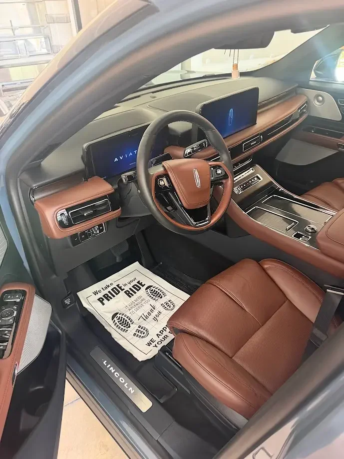Interior of a Lincoln vehicle showcasing a dashboard with two wide digital displays, a steering wheel with a brown leather cover, brown leather seats, and a paper floor mat with footprints and congratulatory message.