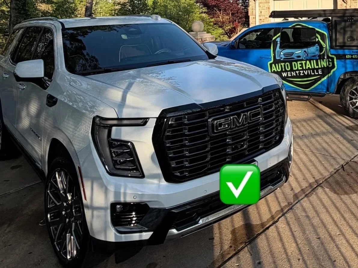 White GMC SUV parked next to a blue vehicle with a sign that reads 'Auto Detailing Wentzville' in a driveway with trees and a house in the background.