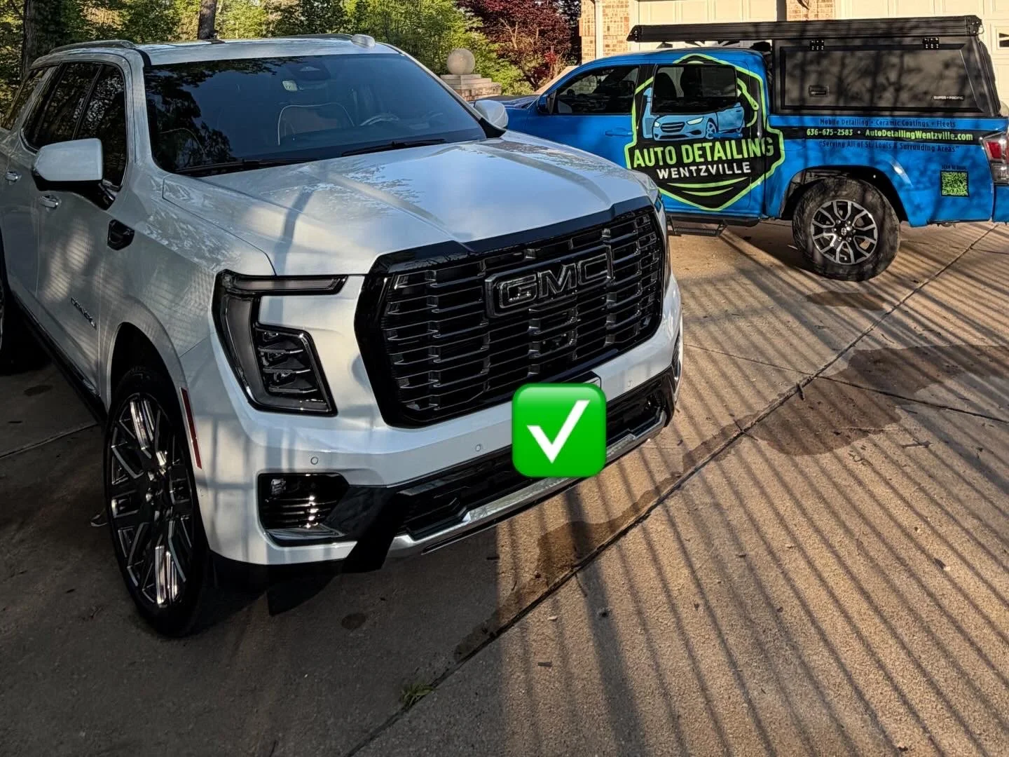 A white GMC SUV parked next to a blue pickup truck with an auto detailing company logo on the side. The SUV is in the foreground, and the pickup truck is in the background on a concrete driveway with shadows and sunlight.