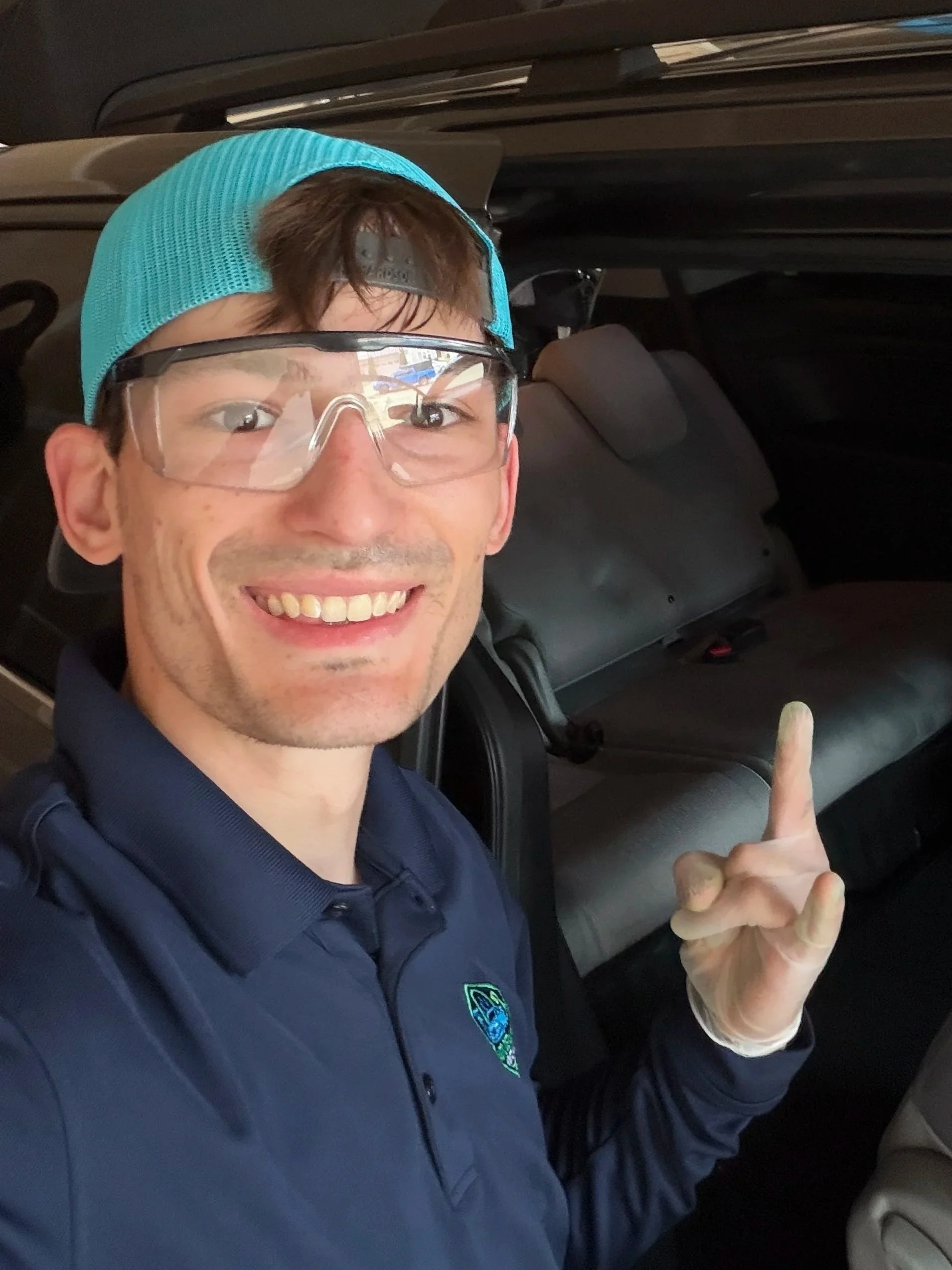 A man wearing a blue cap, safety glasses, and a navy jacket with a police patch is smiling and pointing up with his right index finger while sitting inside a vehicle.