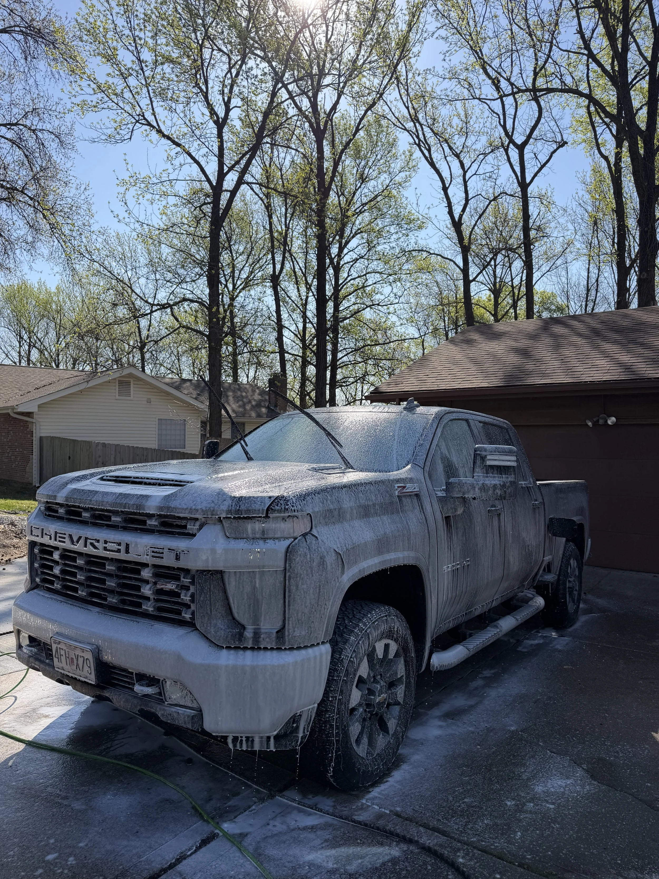 A silver Chevrolet pickup truck covered in soap suds during a car wash in a residential driveway, with houses and tall trees with budding leaves in the background under a clear blue sky.