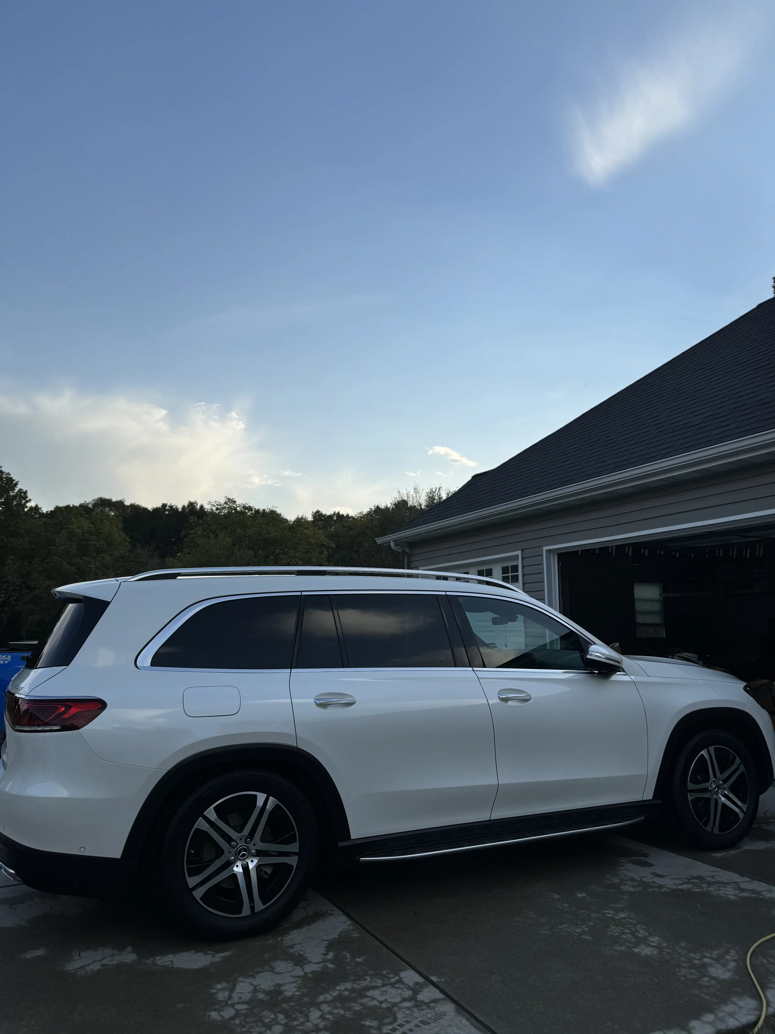 A white SUV parked in a driveway outside a house with a garage. The sky is mostly clear with some clouds in the background.