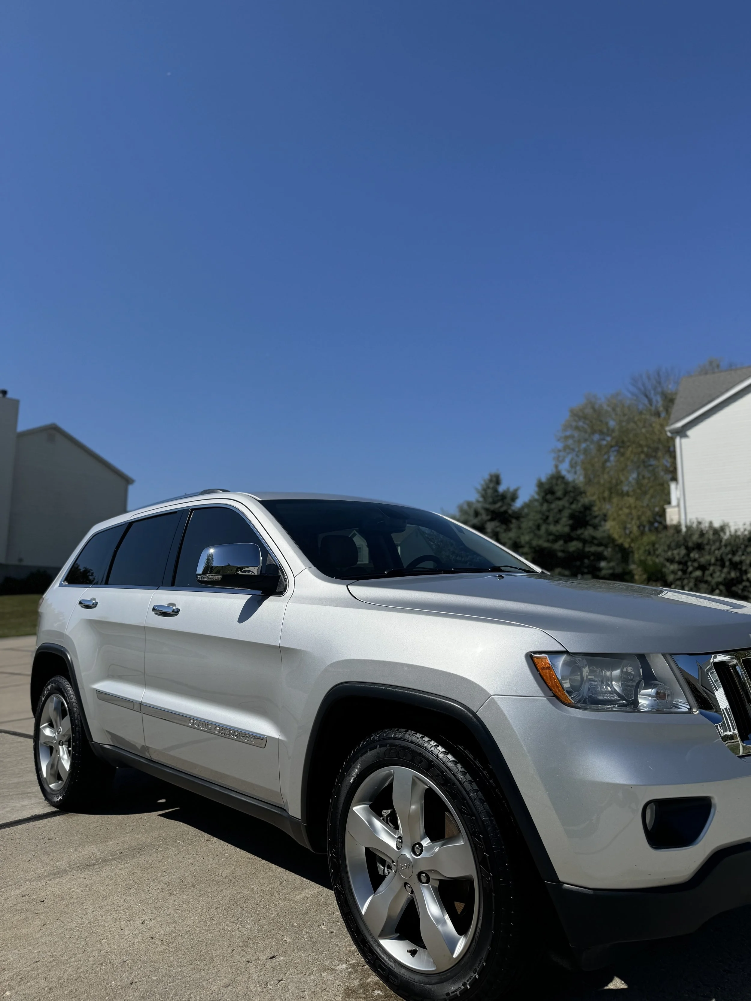 A silver Jeep SUV parked outdoors on a driveway with houses and trees in the background under a clear blue sky.