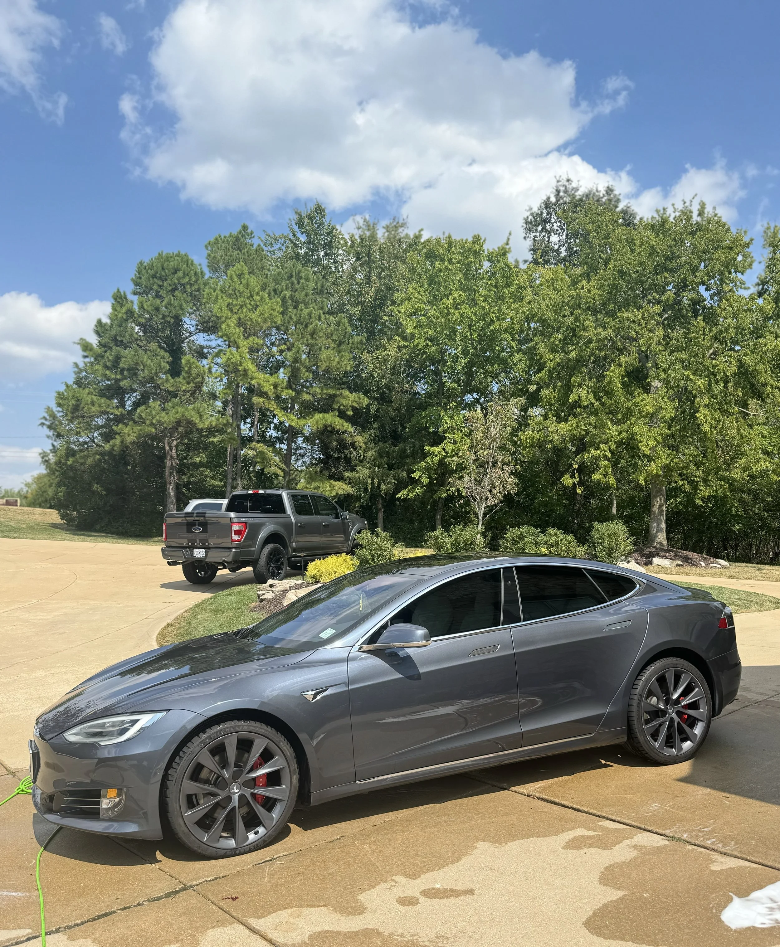 A modern gray Tesla sedan parked on a concrete driveway with a black pickup truck and green trees in the background under a partly cloudy sky.