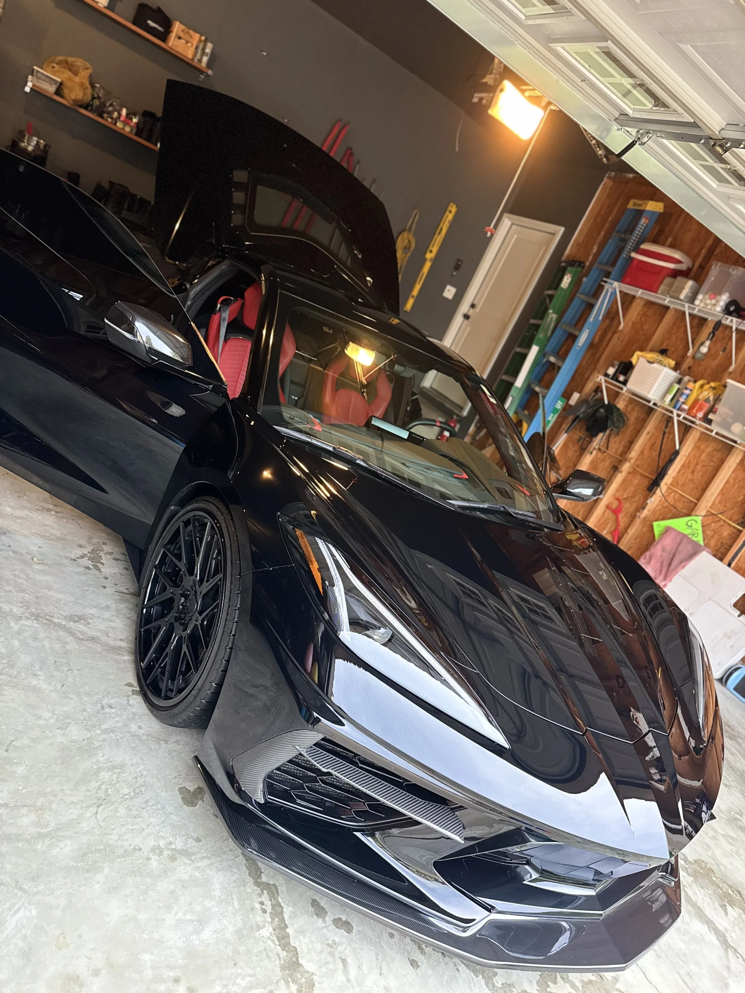 A black luxury sports car with red interior seats parked in a garage. The garage has wooden and gray walls, storage shelves, and tools visible in the background. The car has sleek design details and large black wheels.