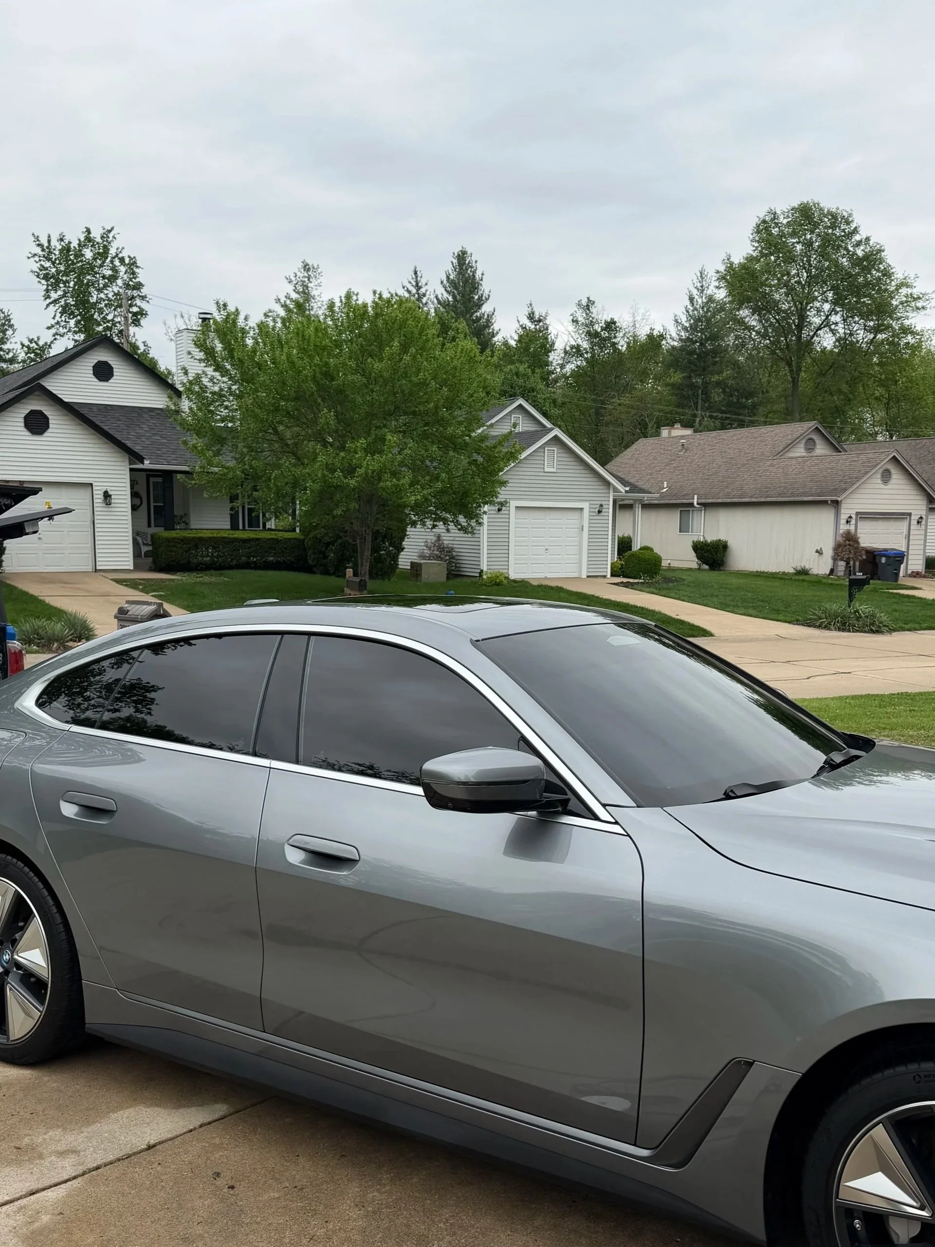 Silver BMW car parked on a driveway in a suburban neighborhood with houses, trees, and an overcast sky in the background.