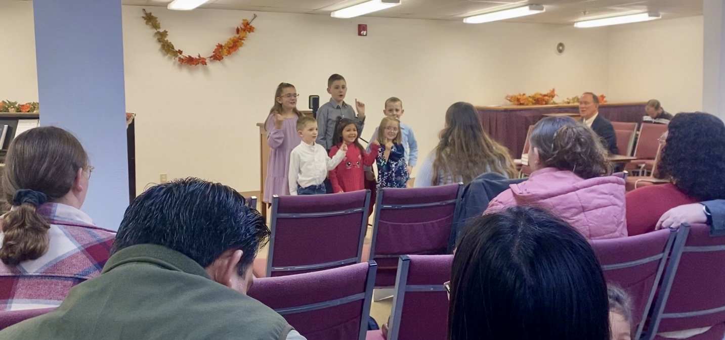 Children performing in a small indoor gathering with an audience seated on maroon chairs, decorations with autumn leaves.