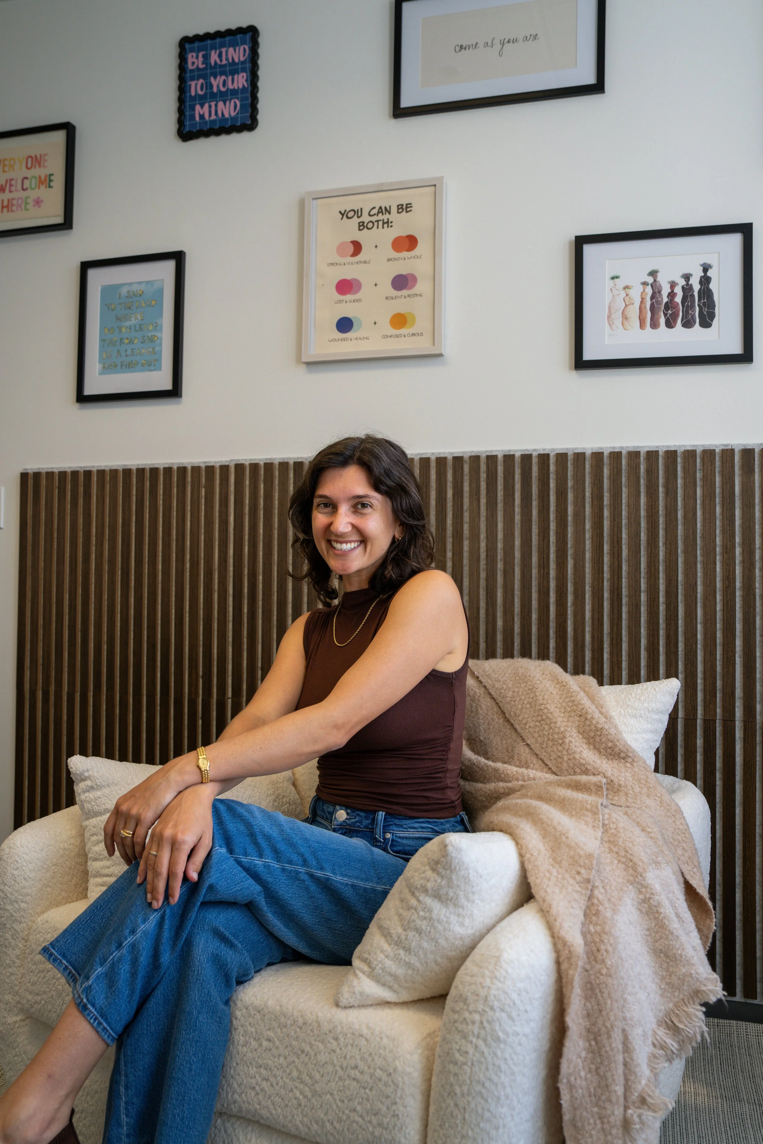 A woman smiling and sitting on a beige armchair with a light brown blanket in a room with framed artwork and motivational quotes on the wall.