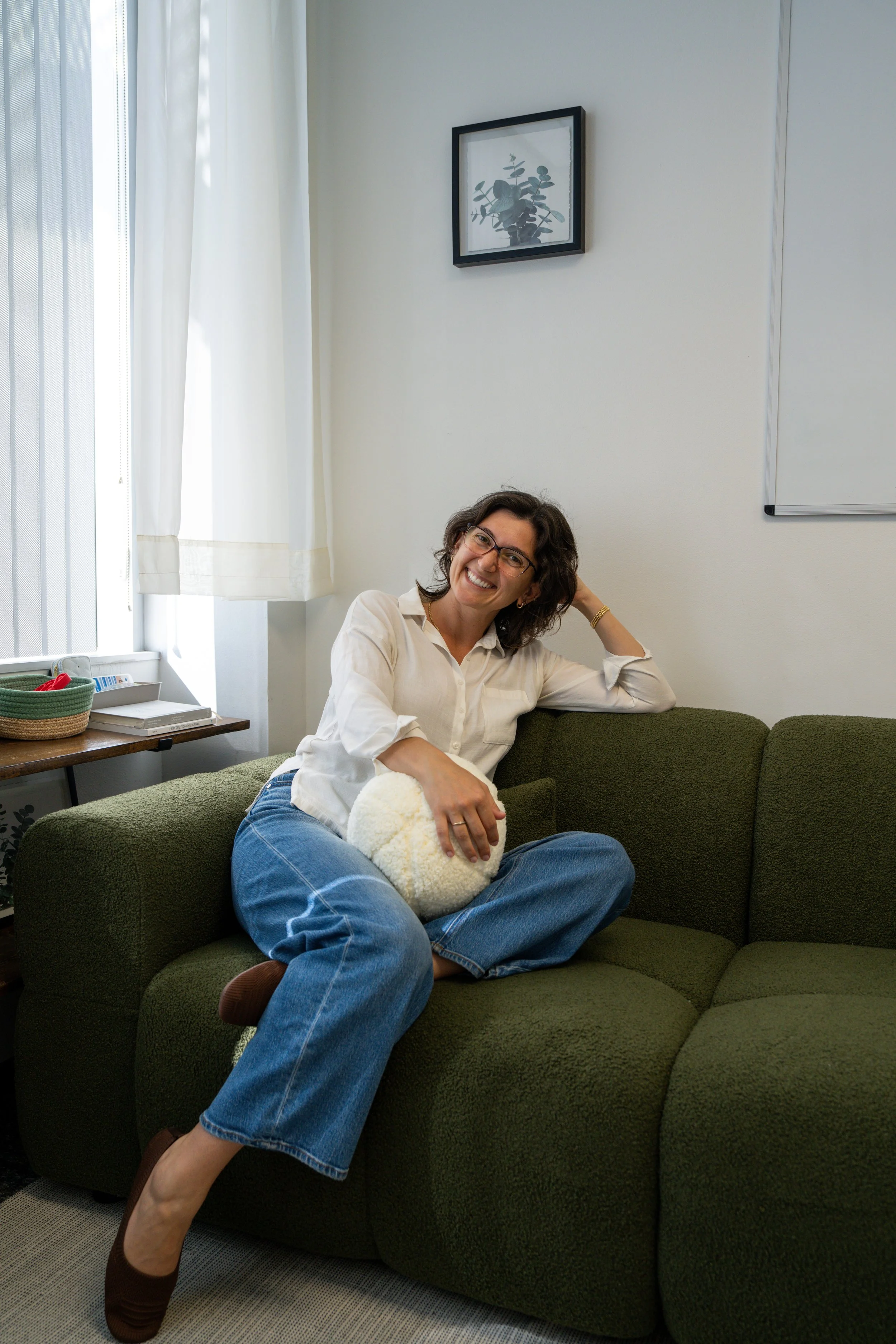 A woman sitting on a green sofa in a bright room, holding a fluffy white pillow, smiling, with a white curtain and framed picture on the wall behind her.