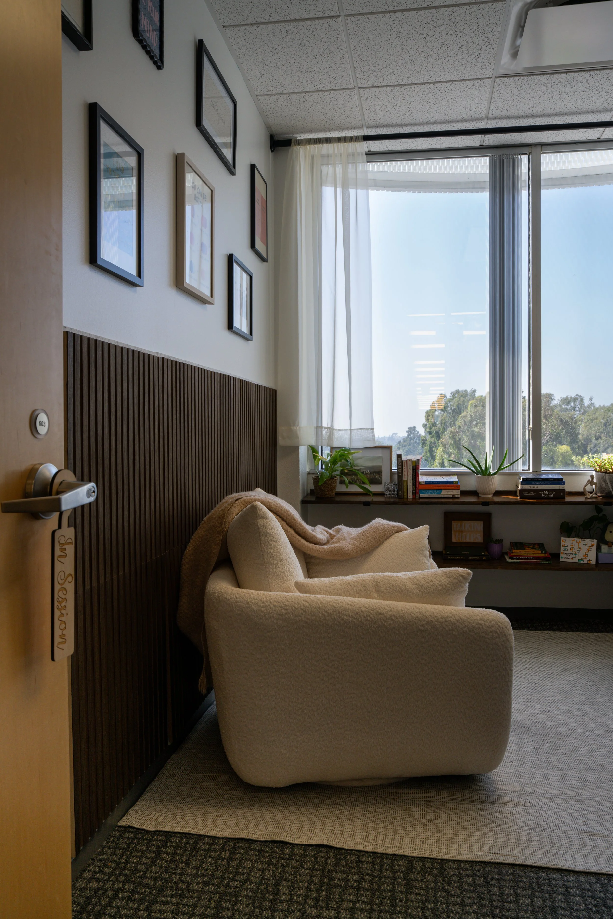 Cozy room with cream armchair, window with curtains, wall decorated with picture frames, bookshelf with plants and books, and outside view of trees during daytime.