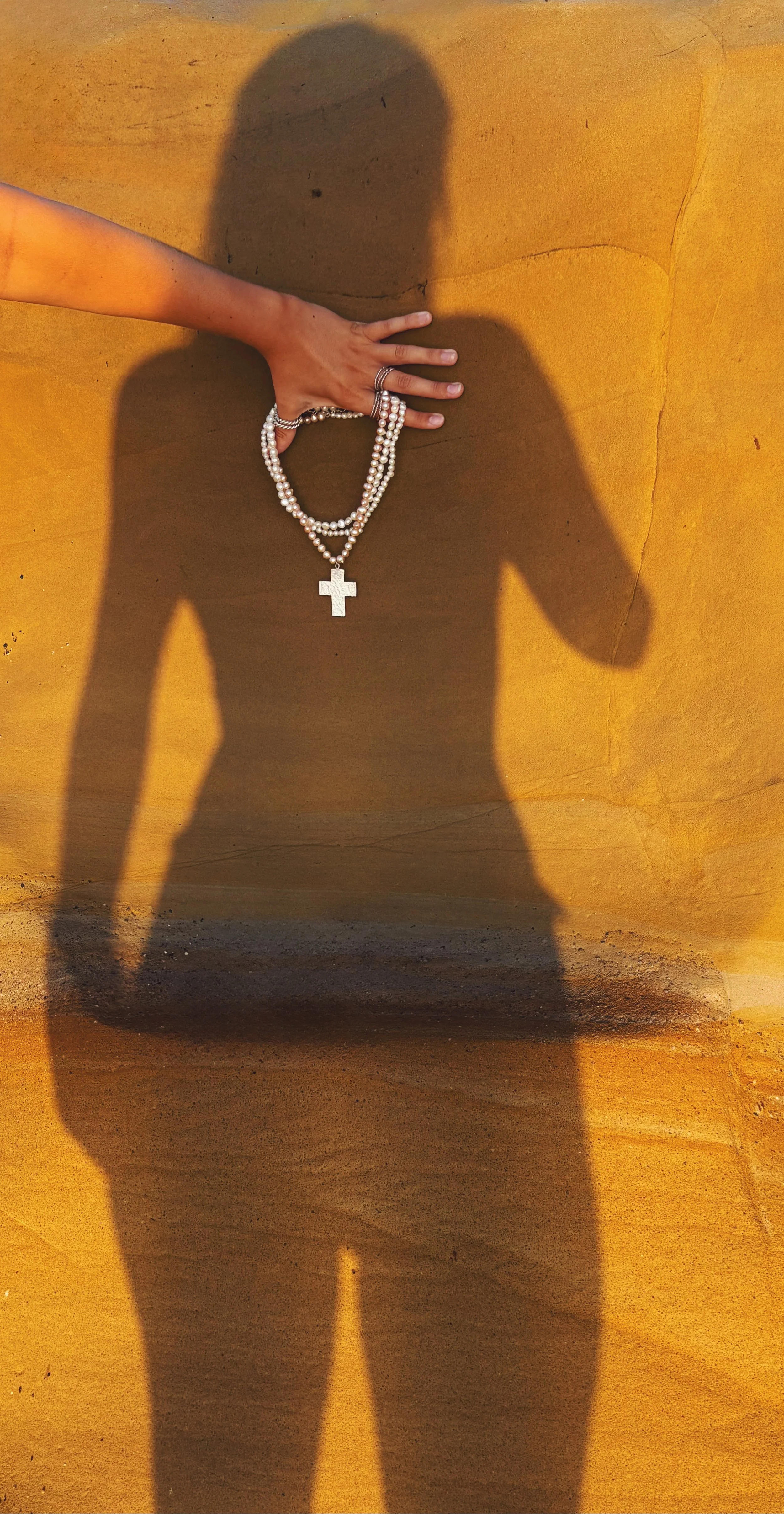 Shadow of a woman holding pearl necklaces with a cross pendant against a yellow wall.