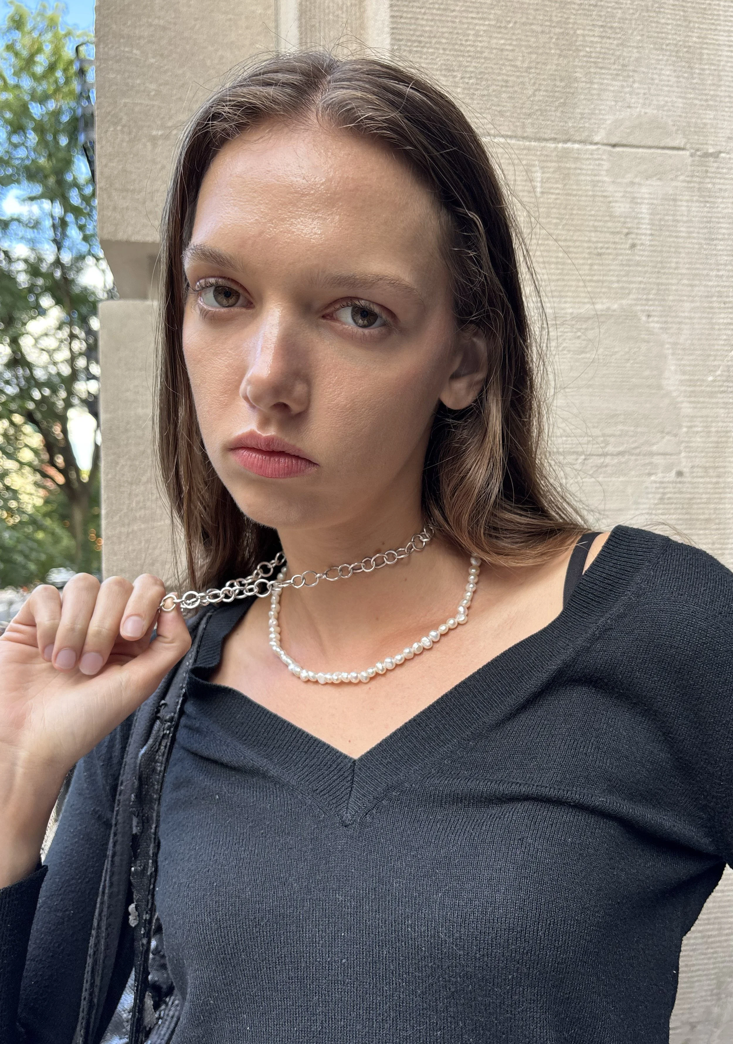 A young woman with light skin, straight brown hair, and gray eyes looking directly at the camera. She is wearing a black top, a pearl necklace, and a silver chain necklace. She is holding the silver chain with her right hand, standing outdoors near a