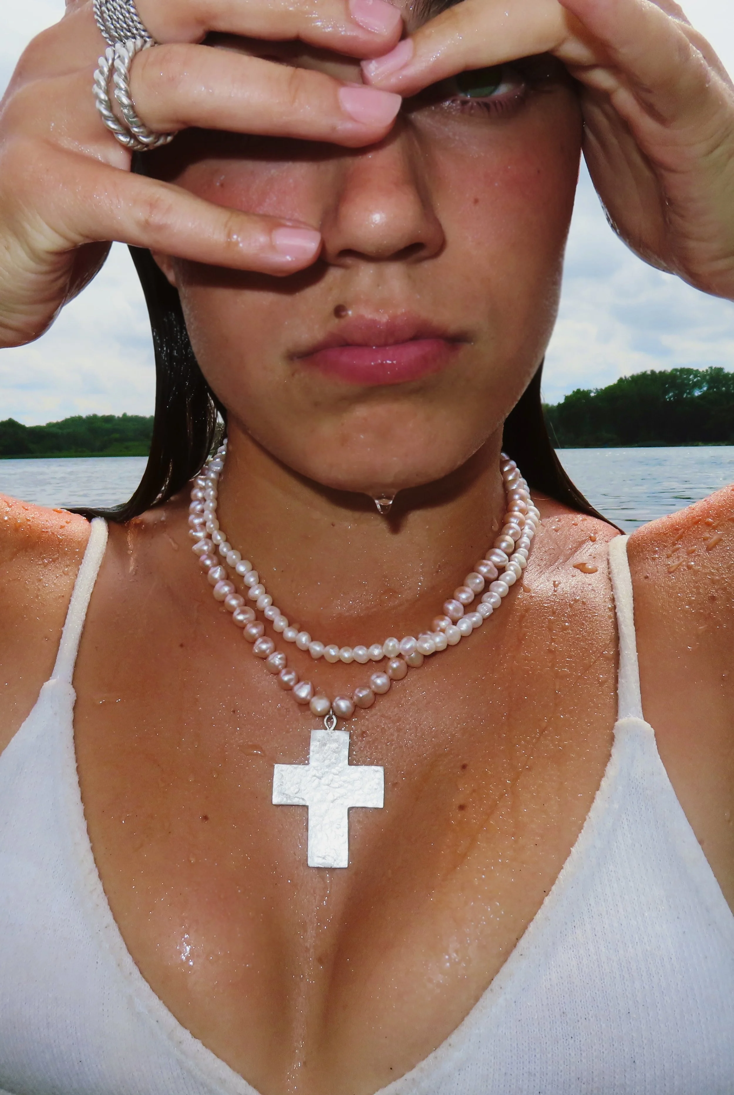 Close-up of a woman wearing pearl necklaces, a white top, and seen partly through her hand with water droplets on her skin, standing near a body of water with a tree-lined shoreline in the background.