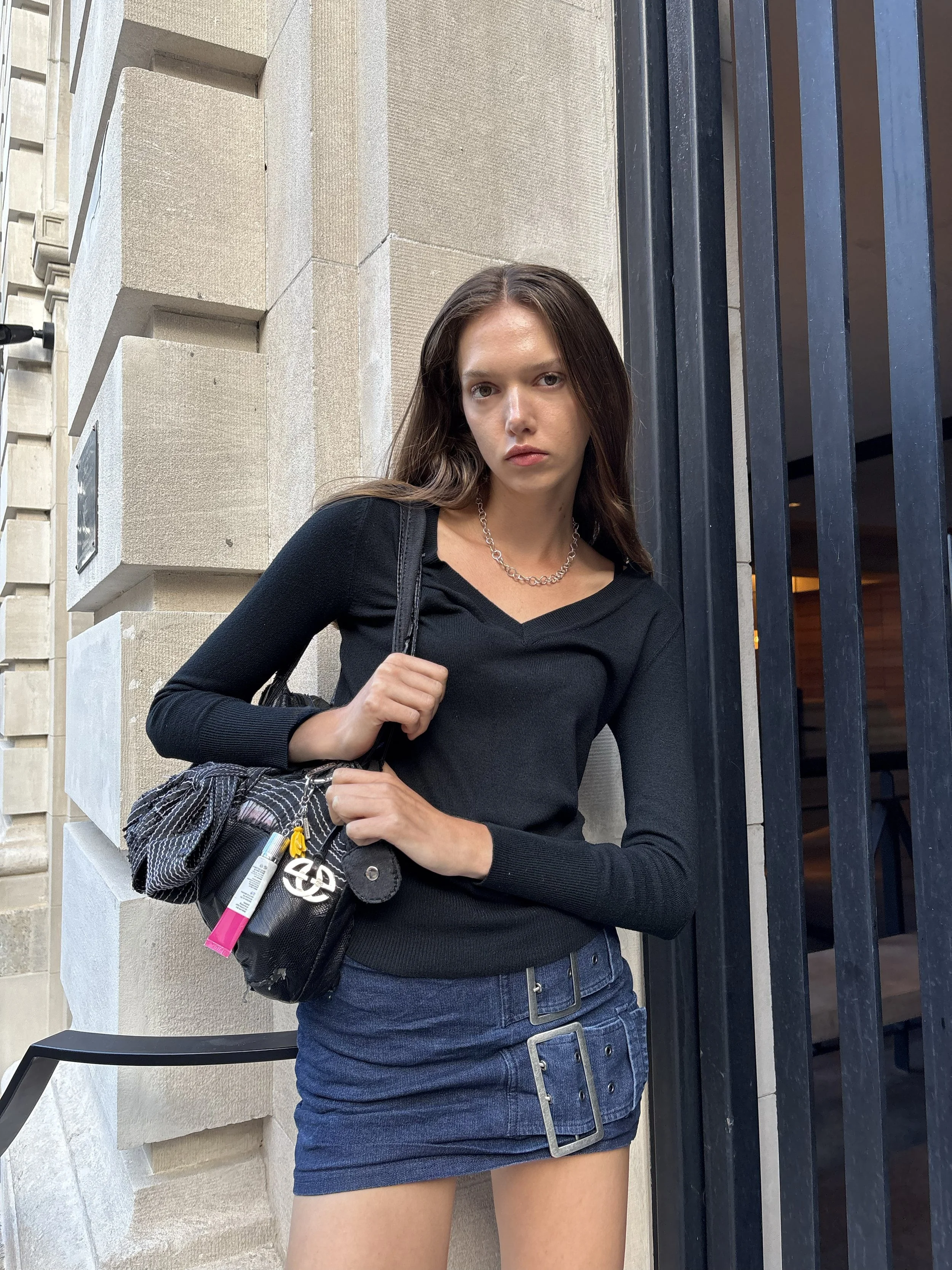 Young woman standing outdoors next to a building with metal gate, wearing a black long-sleeve top, blue denim skirt with buckle details, and carrying a black shoulder bag with a scarf and keychain, looking at the camera.
