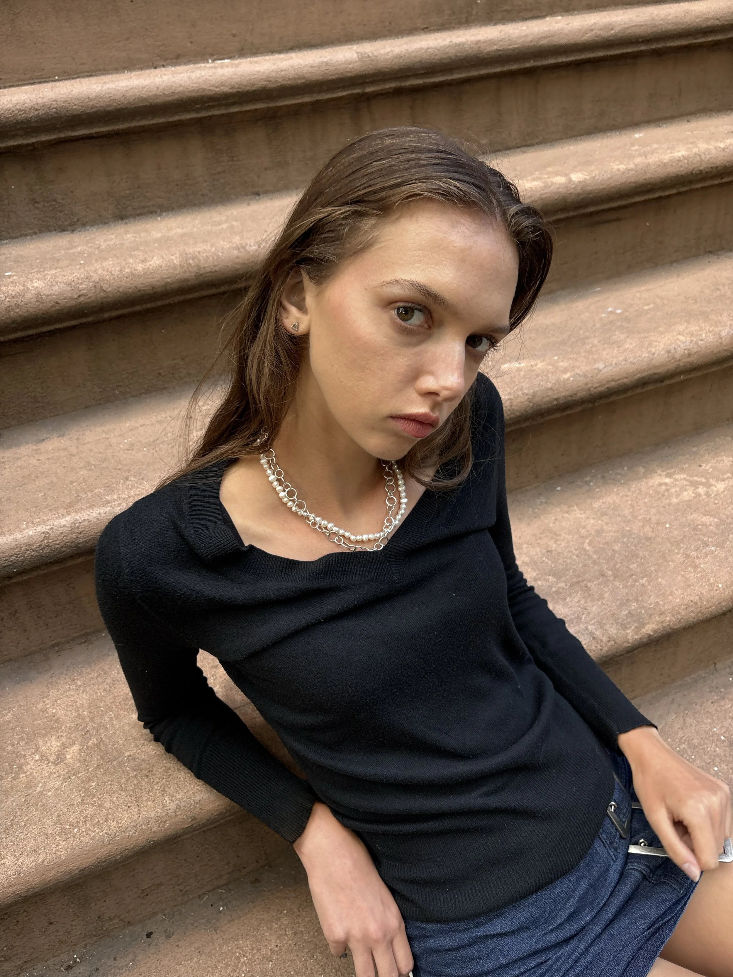A young woman with brown hair sitting onSteps of a brownstone building, wearing a black sweater, blue jeans, and a pearl necklace, looking at the camera.
