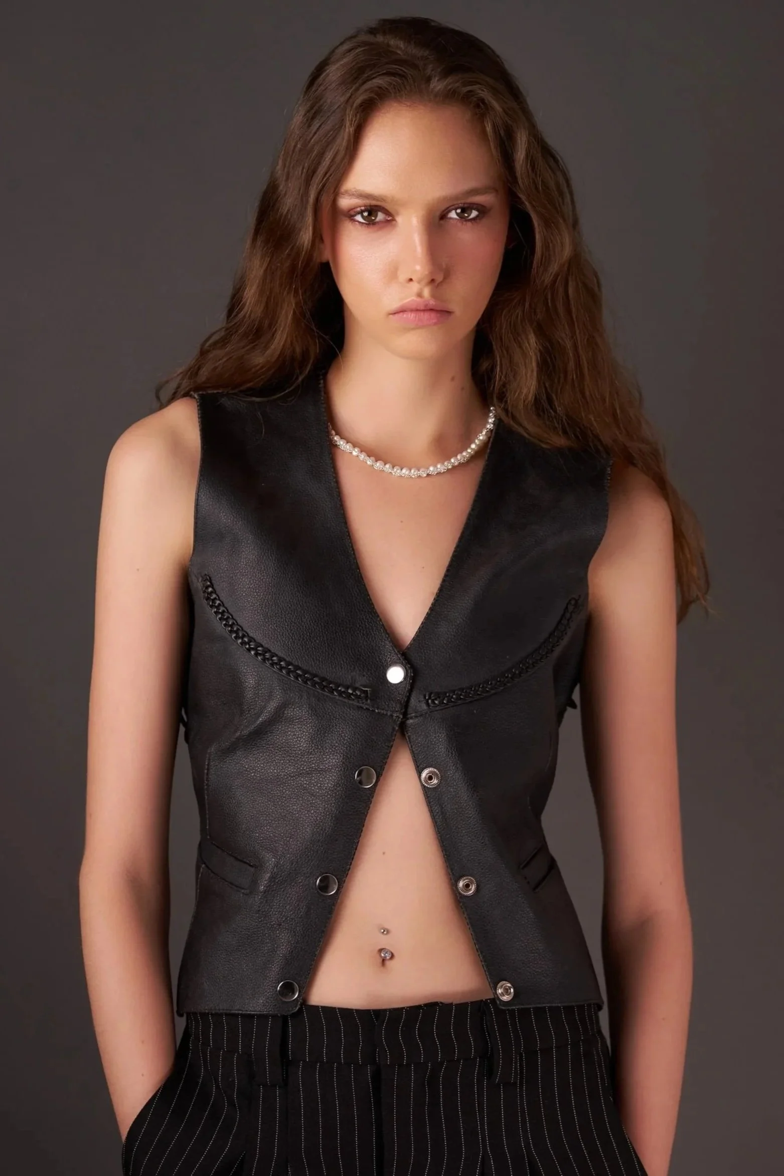 A young woman with long wavy brown hair and piercing eyes, wearing a sleeveless black leather vest with metallic details, a pearl necklace, and black pinstriped pants, standing against a dark background.