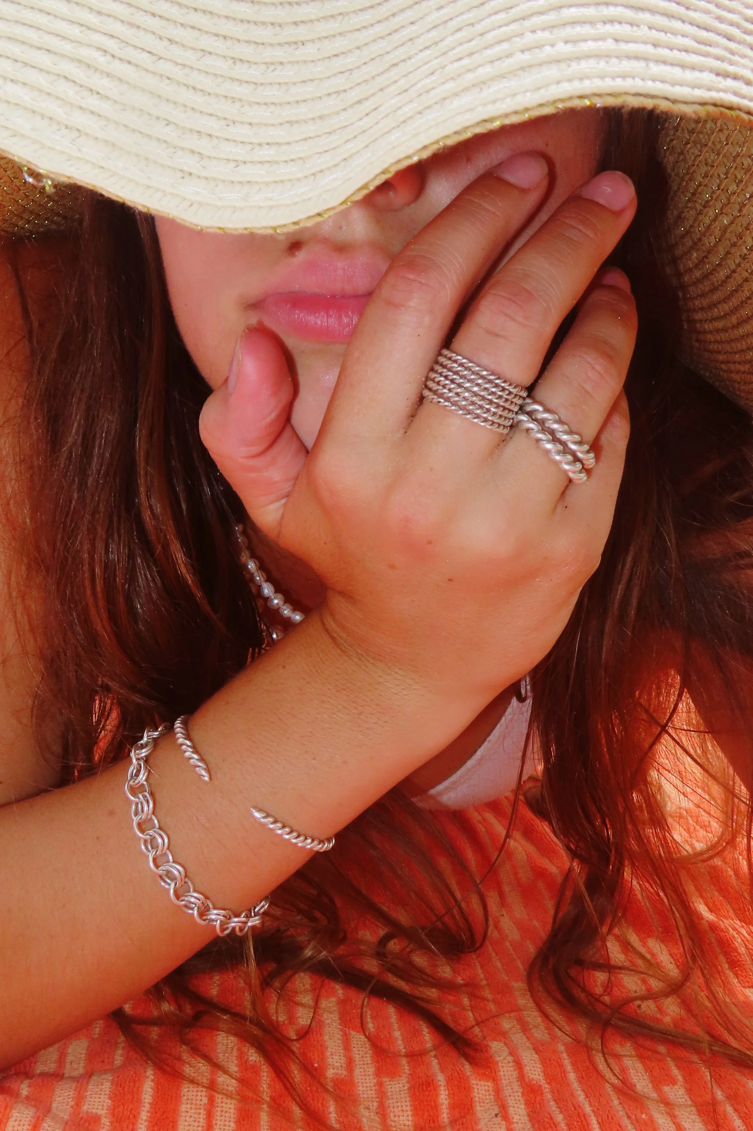 A woman with brown hair partially covered by a wide-brimmed straw hat, wearing several silver rings and bracelets, resting her chin on her hand with her face partially obscured by the hat.
