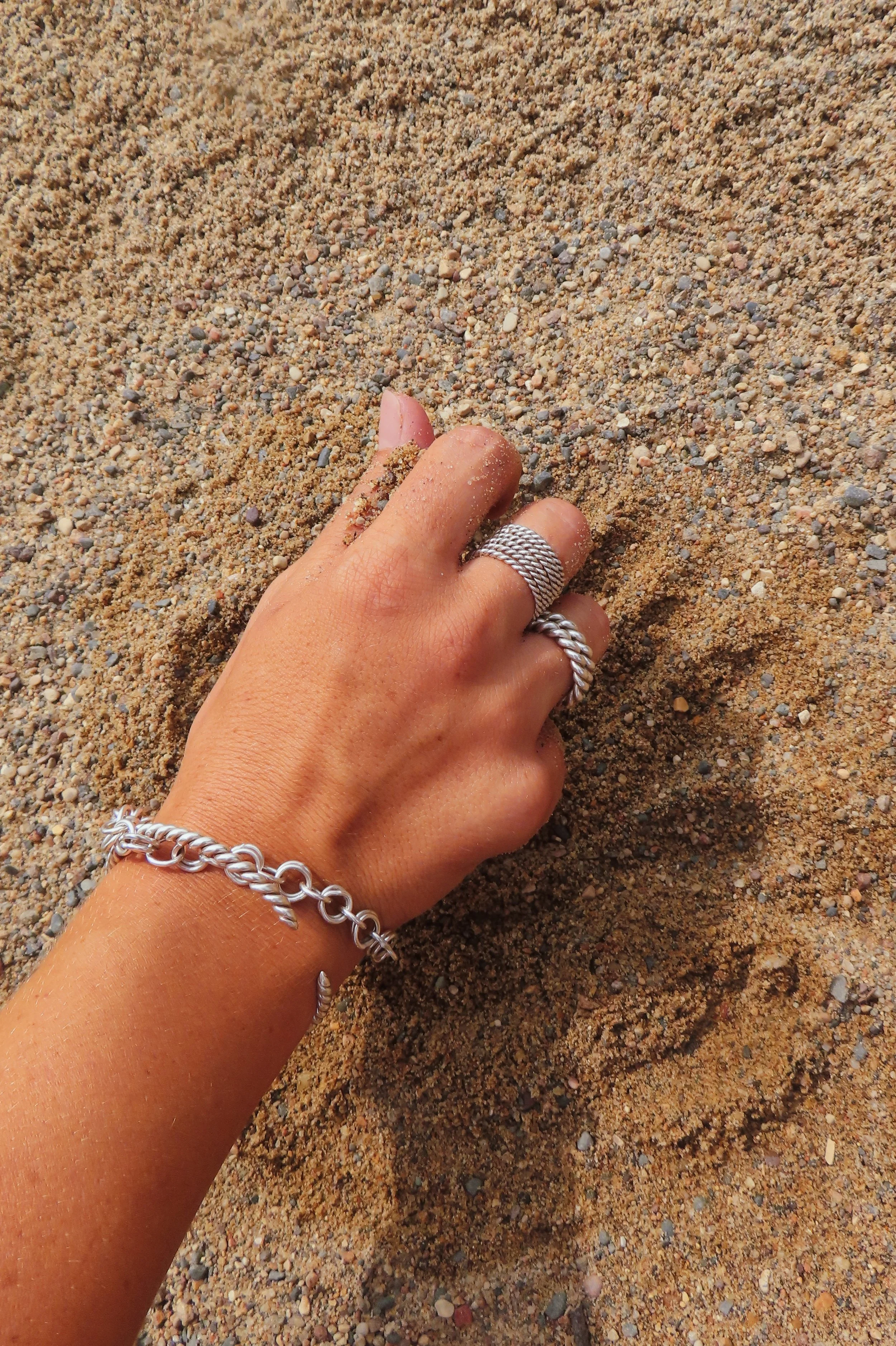 A hand with jewelry, including rings and a bracelet, partially buried in sandy beach with small pebbles.