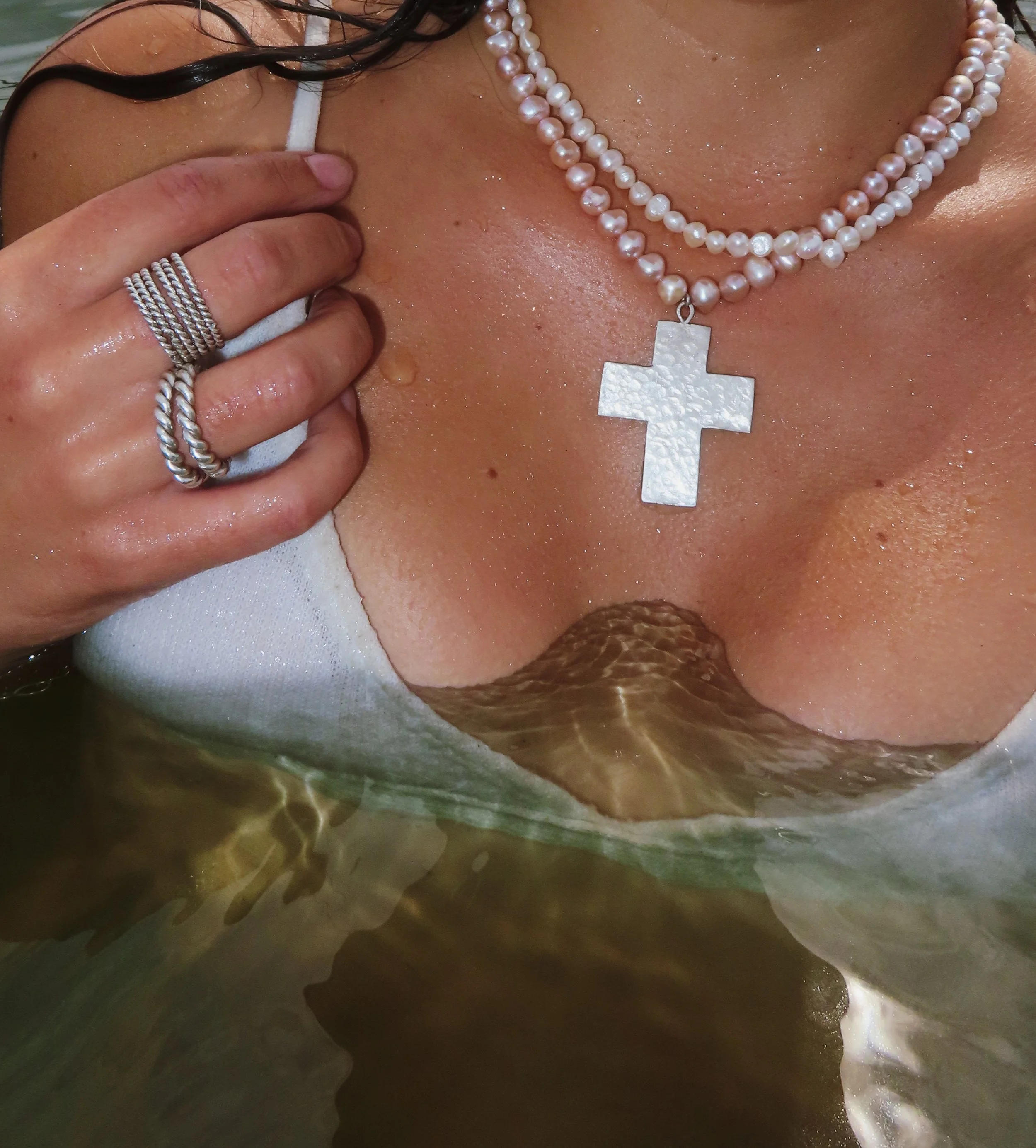 A woman wearing pearl necklaces, rings, and a cross pendant, partially submerged in water, with wet skin and jewelry.