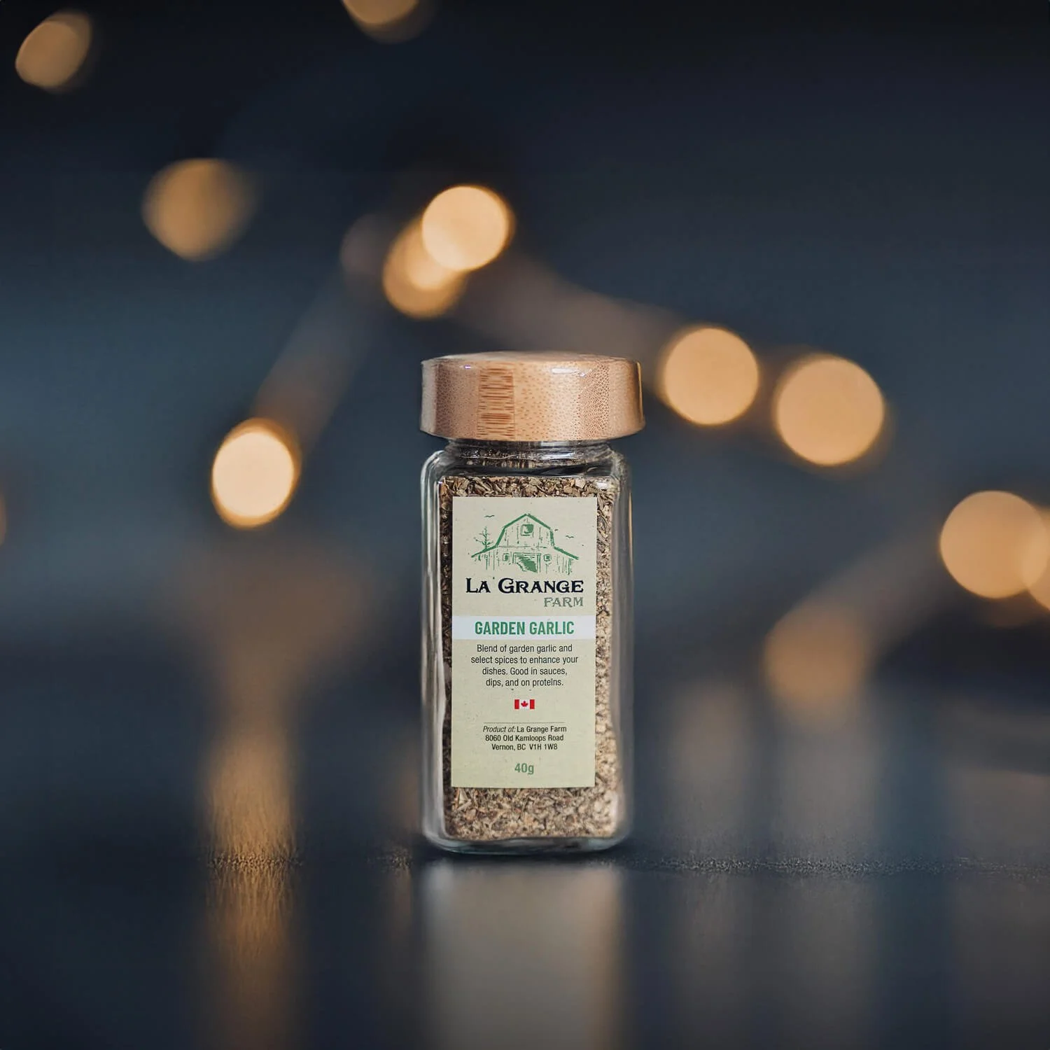 Glass jar of La Grange Farm garden garlic seasoning on a dark surface with blurred lights in the background.