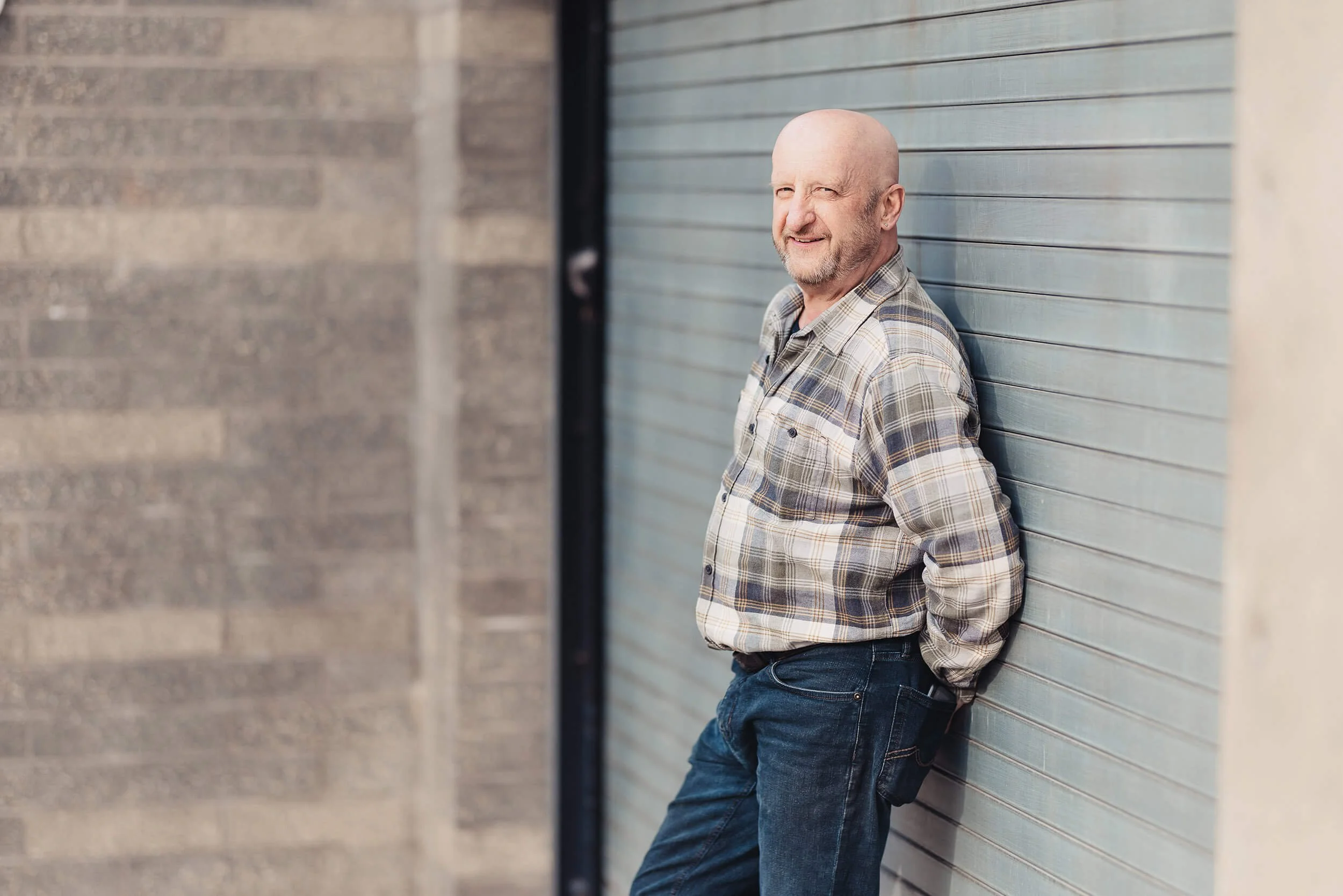 Smiling Kenny in plaid shirt and jeans leaning against a metal garage door with hands in pockets.