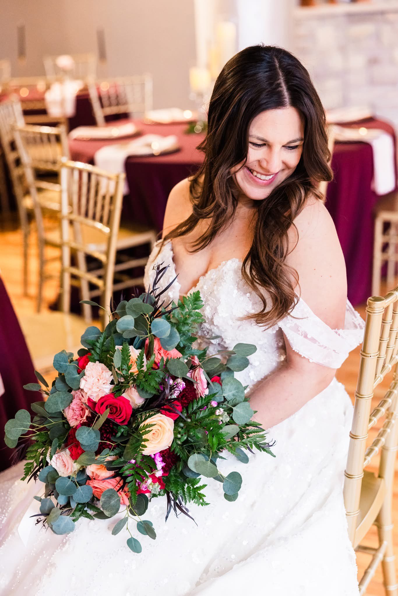 Bride smiling while holding a colorful bouquet, wearing a white wedding dress, seated in a room with tables and gold chairs.