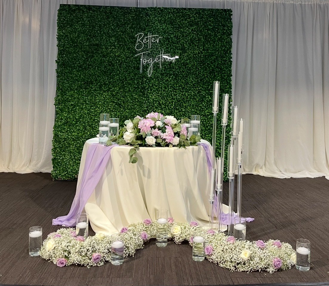 Wedding backdrop with a green wall and 'Better Together' sign, adorned with floral arrangements and candles.