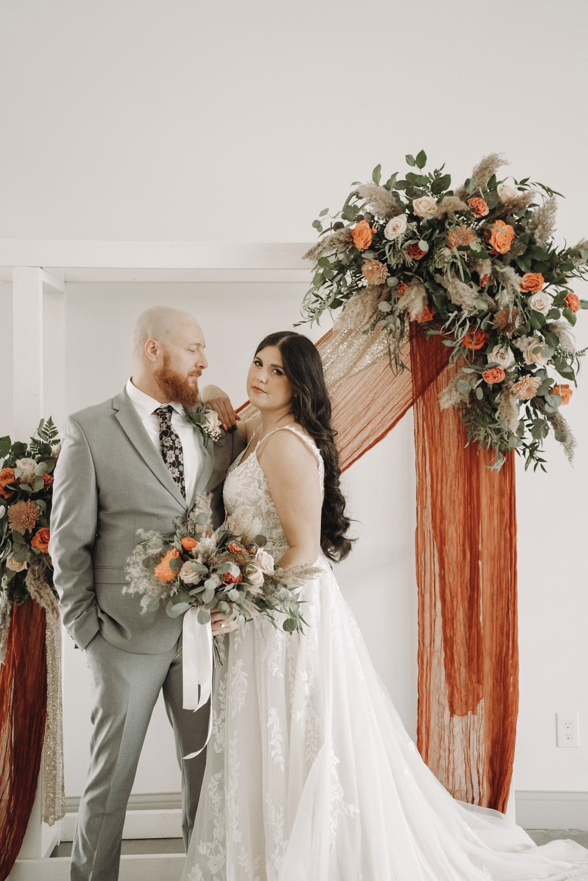 Bride and groom posing in elegant attire next to a floral arch with orange and white flowers, dressed in wedding gown and suit, holding bouquets.
