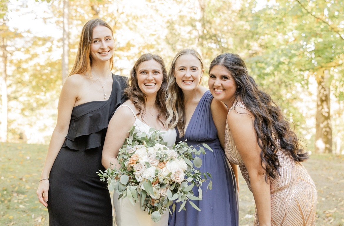 Four women posing outdoors, with the central woman wearing a white dress and holding a bouquet. The others are in dresses of black, blue, and gold. Trees with green leaves are in the background.
