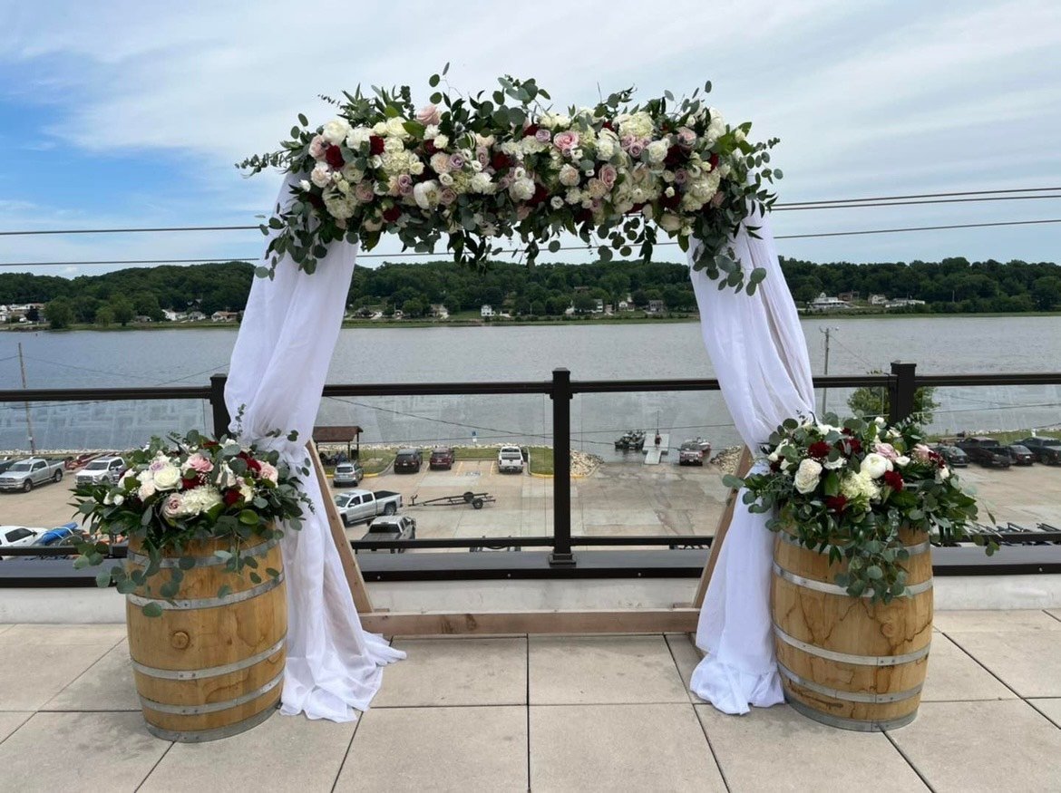 Outdoor wedding arch with floral decorations and white drapery on wooden barrels by a waterfront.