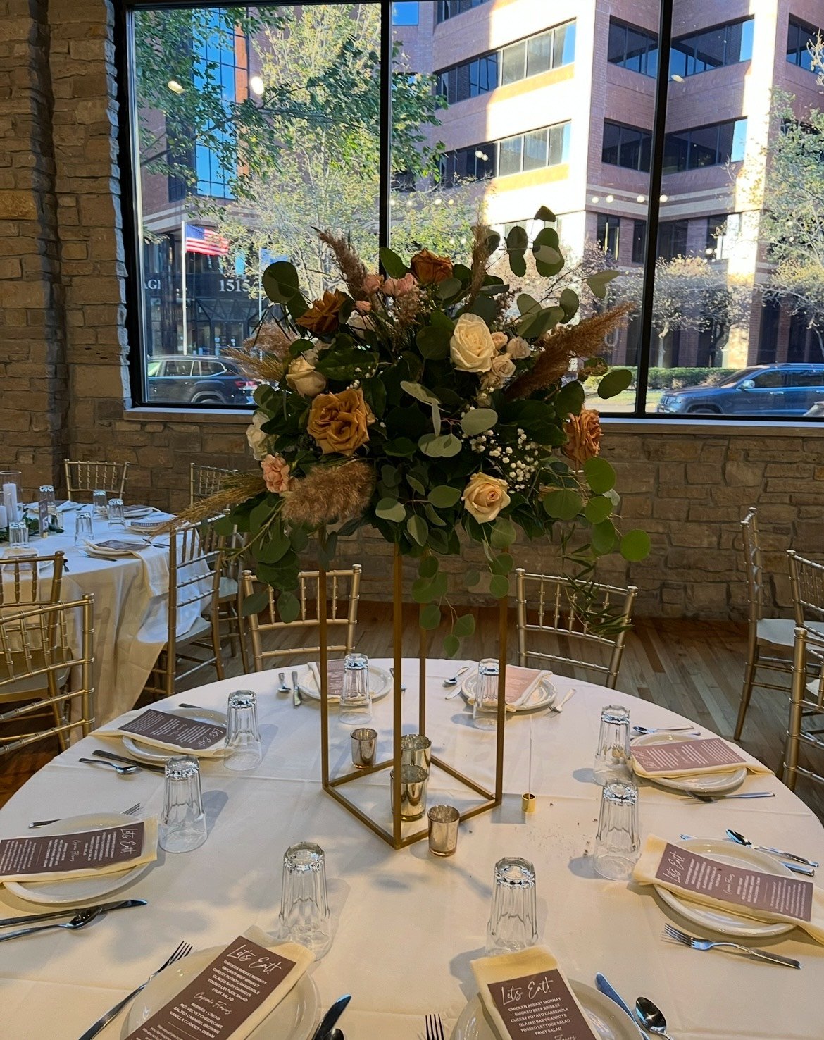 Elegant wedding reception table with floral centerpiece, menu cards, and gold chairs in a room with large windows showing a cityscape.