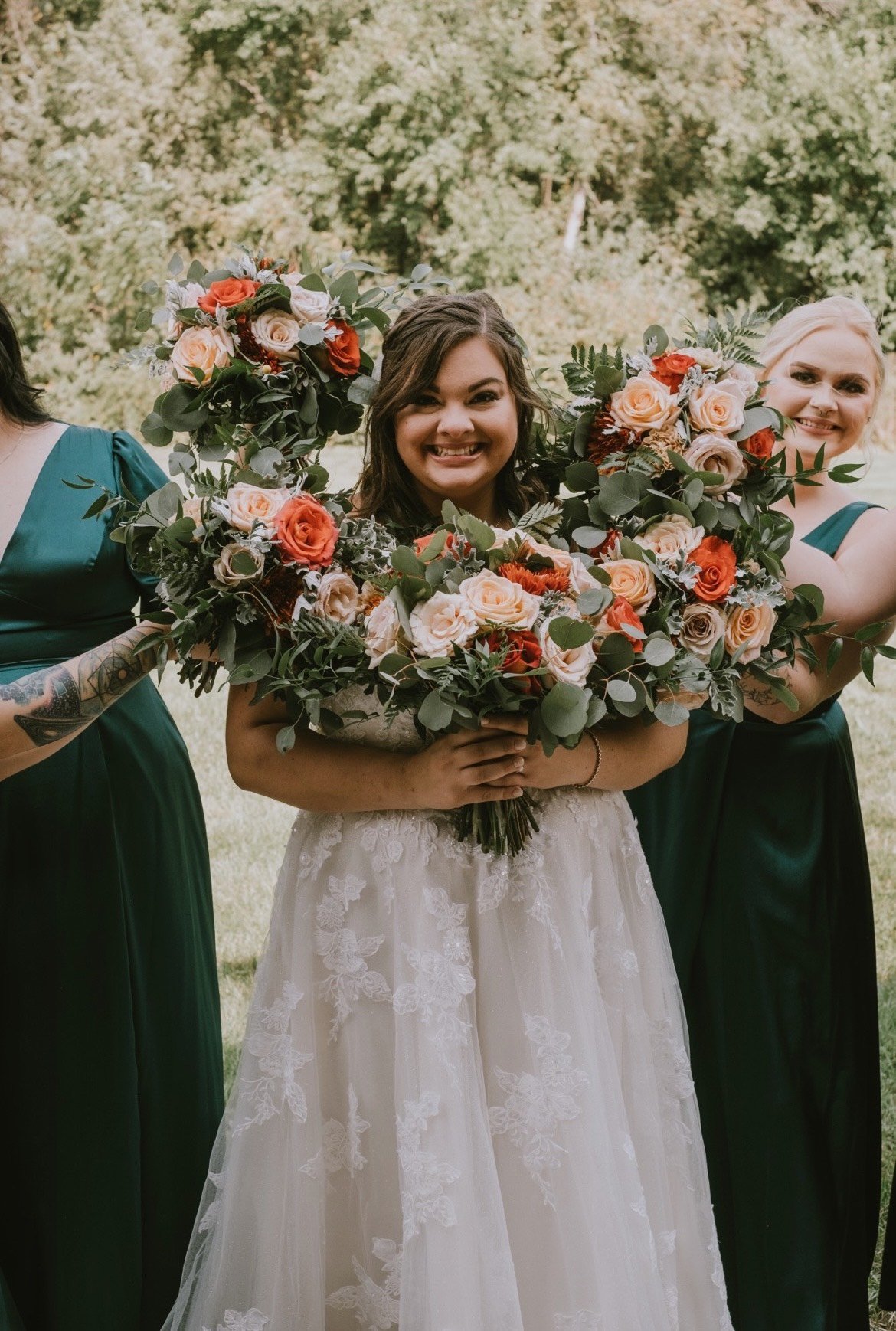 Bride smiling, holding floral bouquets with bridesmaids in green dresses outdoors.