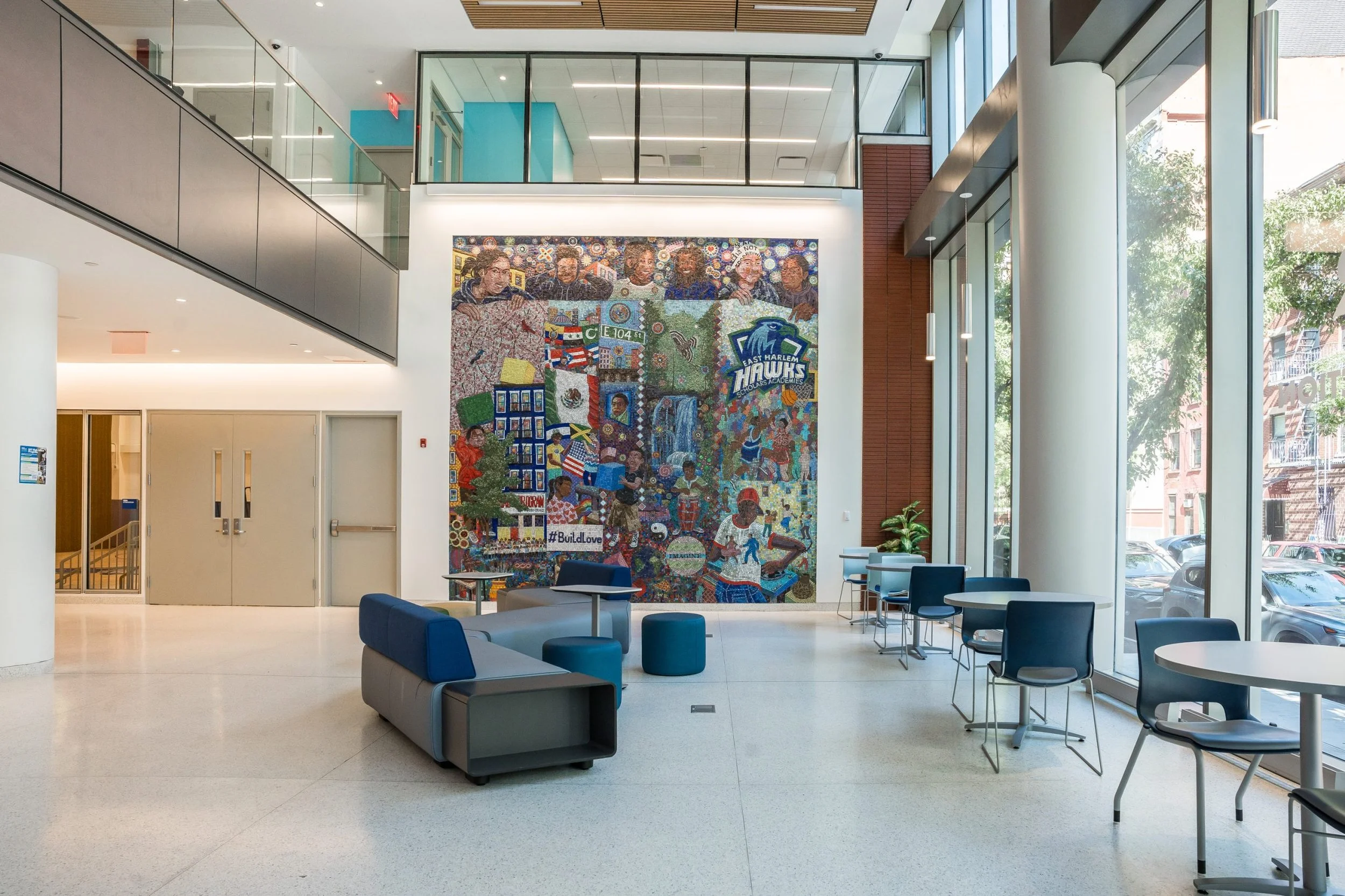 Modern lobby with large colorful mosaic mural, seating area with blue and gray chairs, and floor-to-ceiling windows letting in natural light.