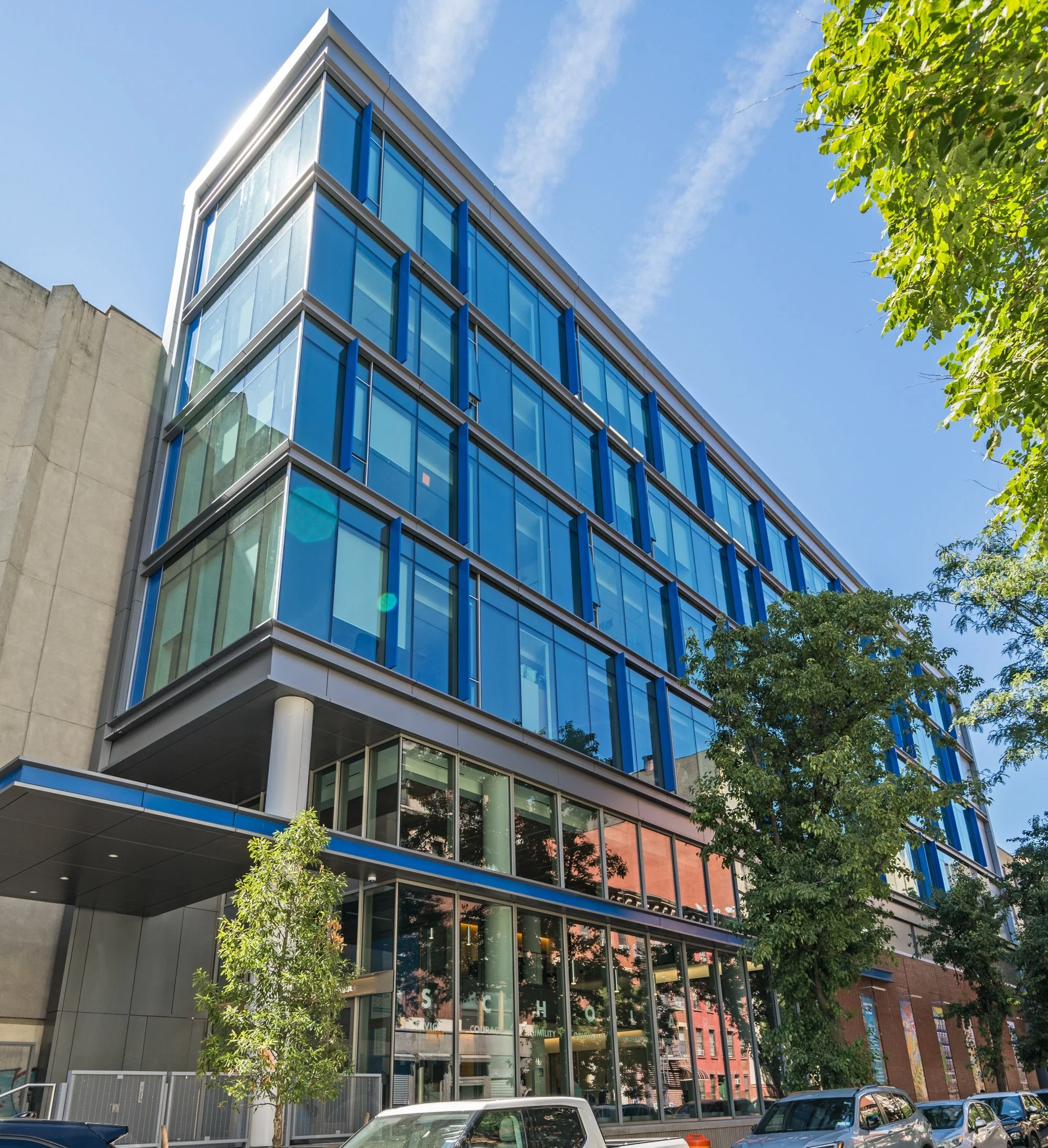 Modern multi-story glass office building with trees and parked cars in front, under a clear blue sky with some clouds.
