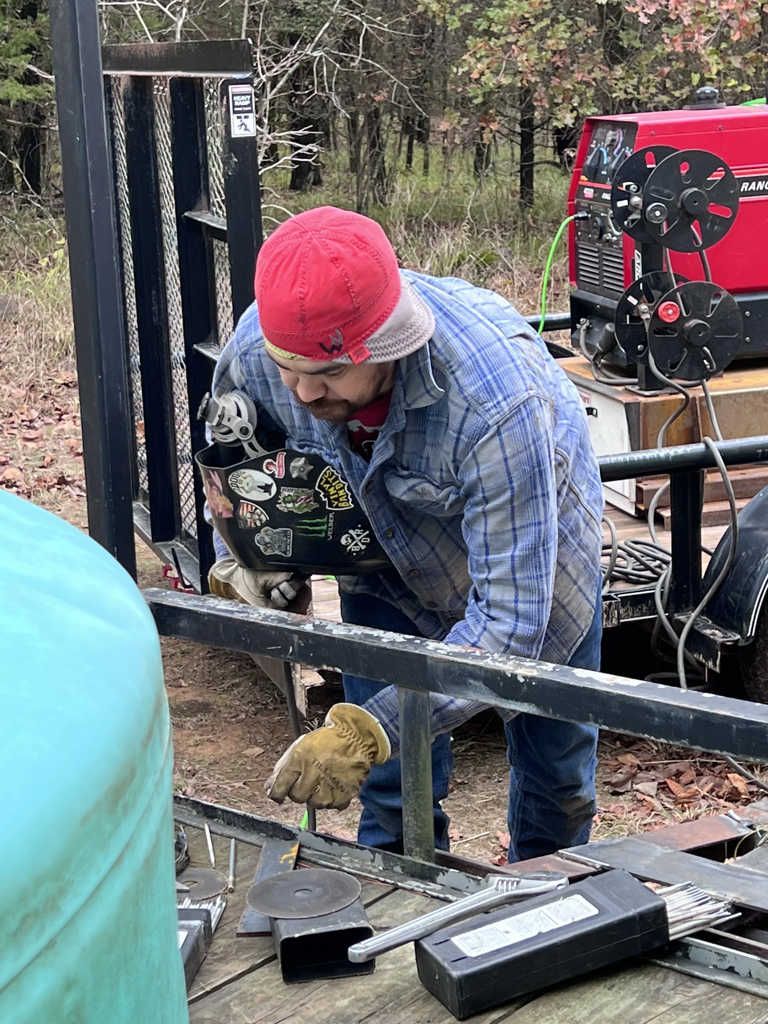 A man wearing a red cap, plaid shirt, and gloves welding outdoors on a trailer in a wooded area.
