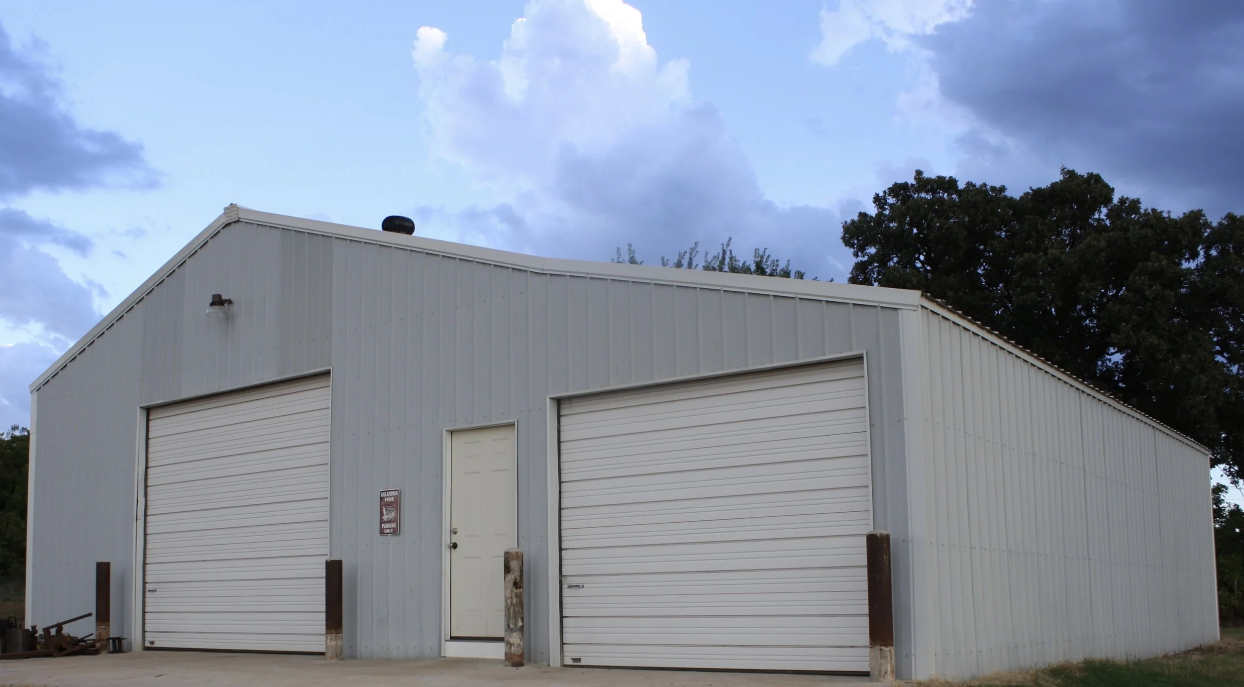 A large light gray metal storage building with two white roll-up garage doors, a narrow white door in the middle, and a sign on the wall near the door. There are two metal posts in front of the building and some equipment on the ground to the left. The sky is partly cloudy with large clouds and some trees in the background.