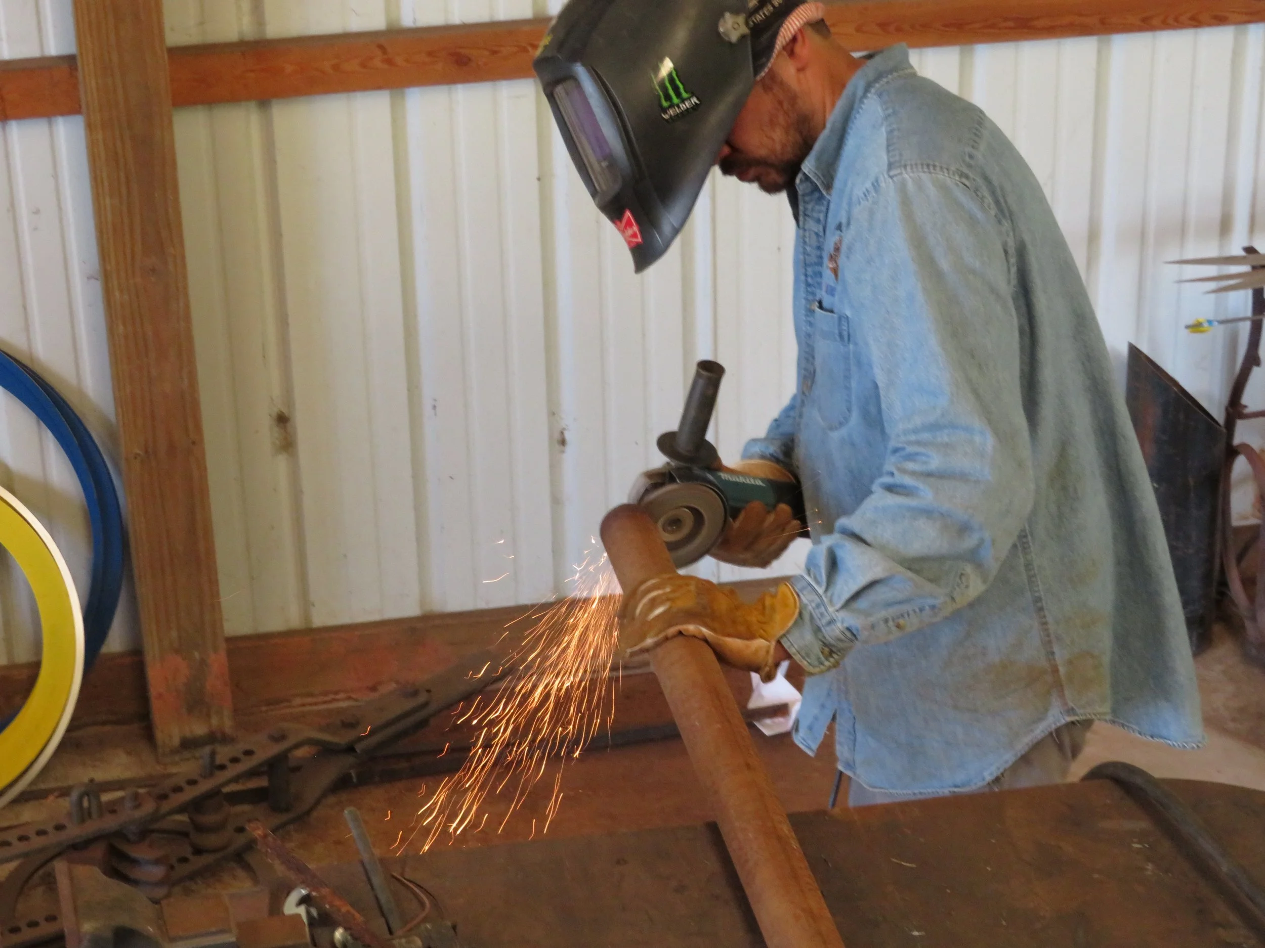 A man wearing a welding helmet and gloves is grinding a metal pipe, creating sparks in a workshop.