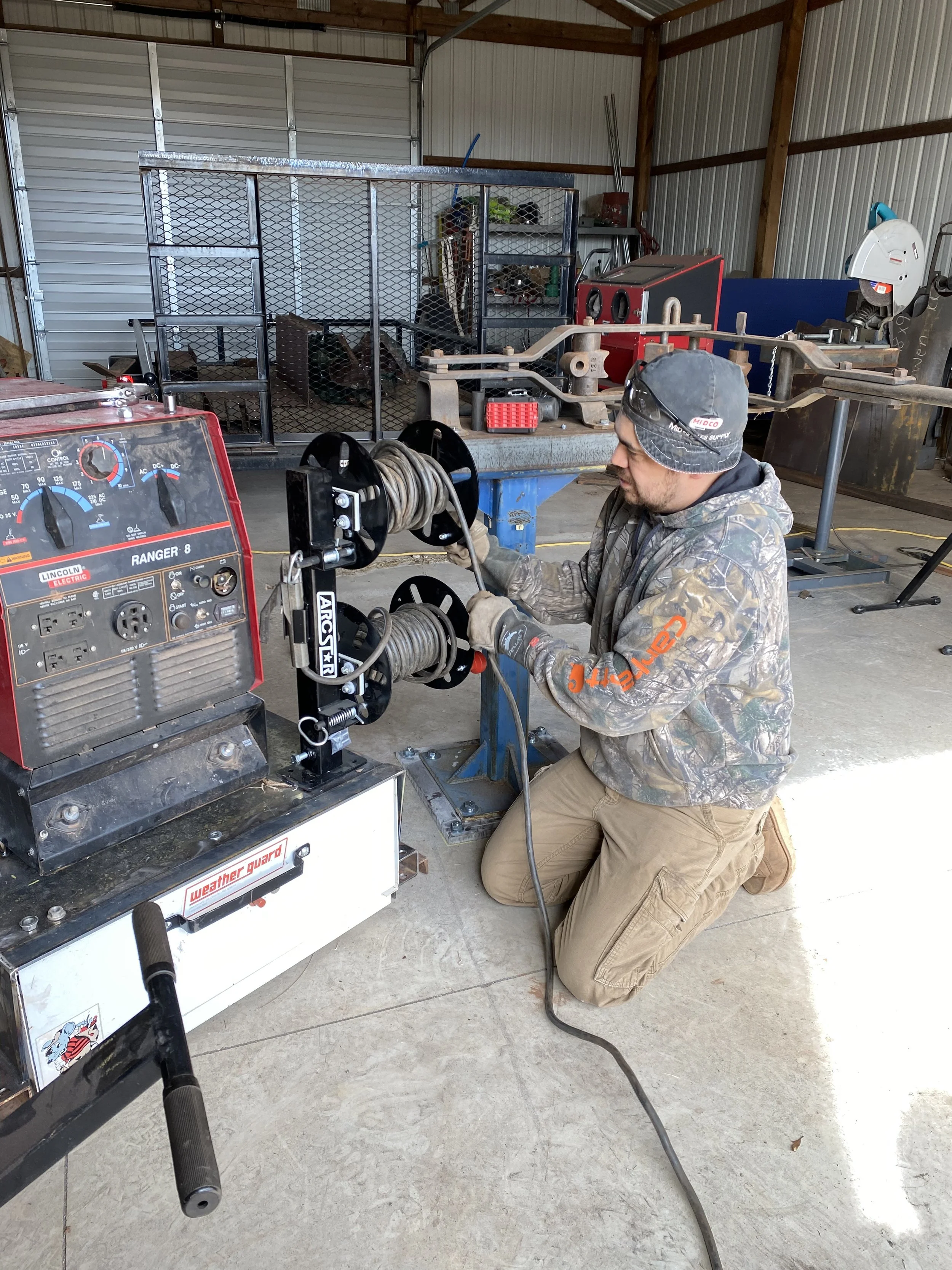 A man kneeling on the concrete floor in a workshop, operating a Lincoln Ranger 8 welding machine and two spool guns with cable reels.