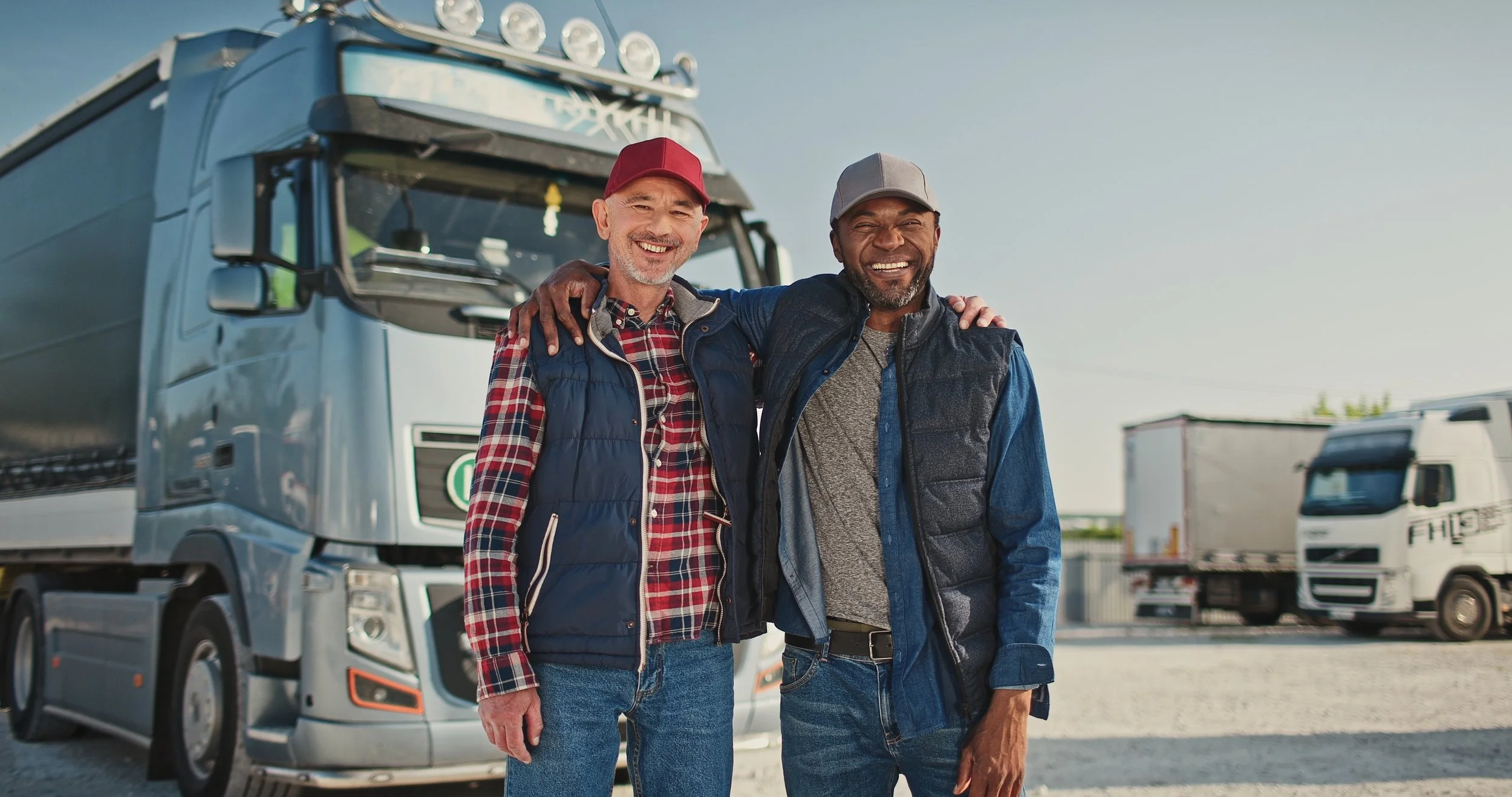 Two smiling men in casual clothing with arms around each other's shoulders, standing in front of moving trucks, outdoors on a sunny day.