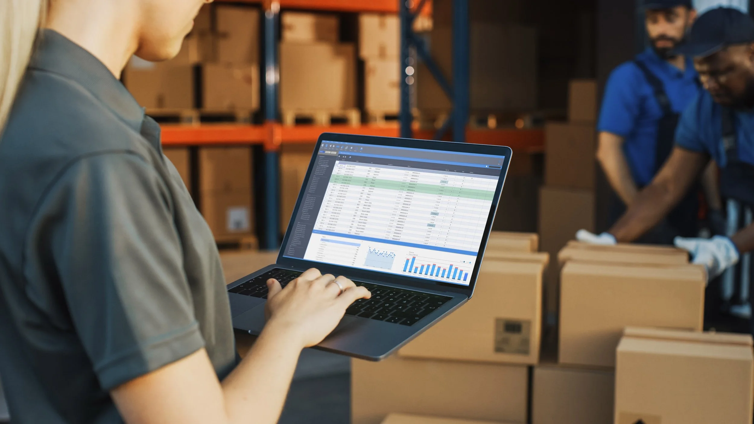 A warehouse worker is viewing a spreadsheet with graphs on a laptop in a storage area with boxes and shelving in the background.