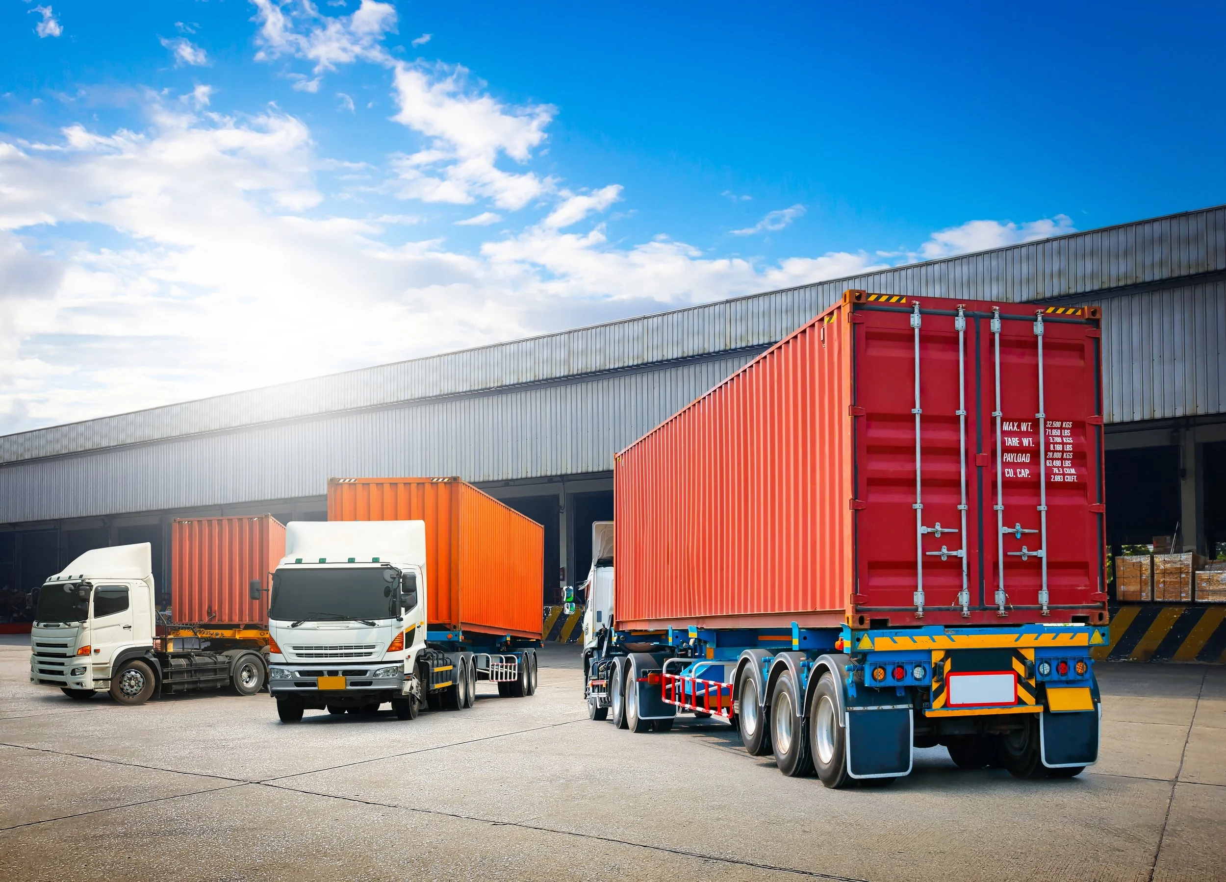 Multiple trucks, including one large red container truck, parked outside a warehouse under a blue sky with clouds.