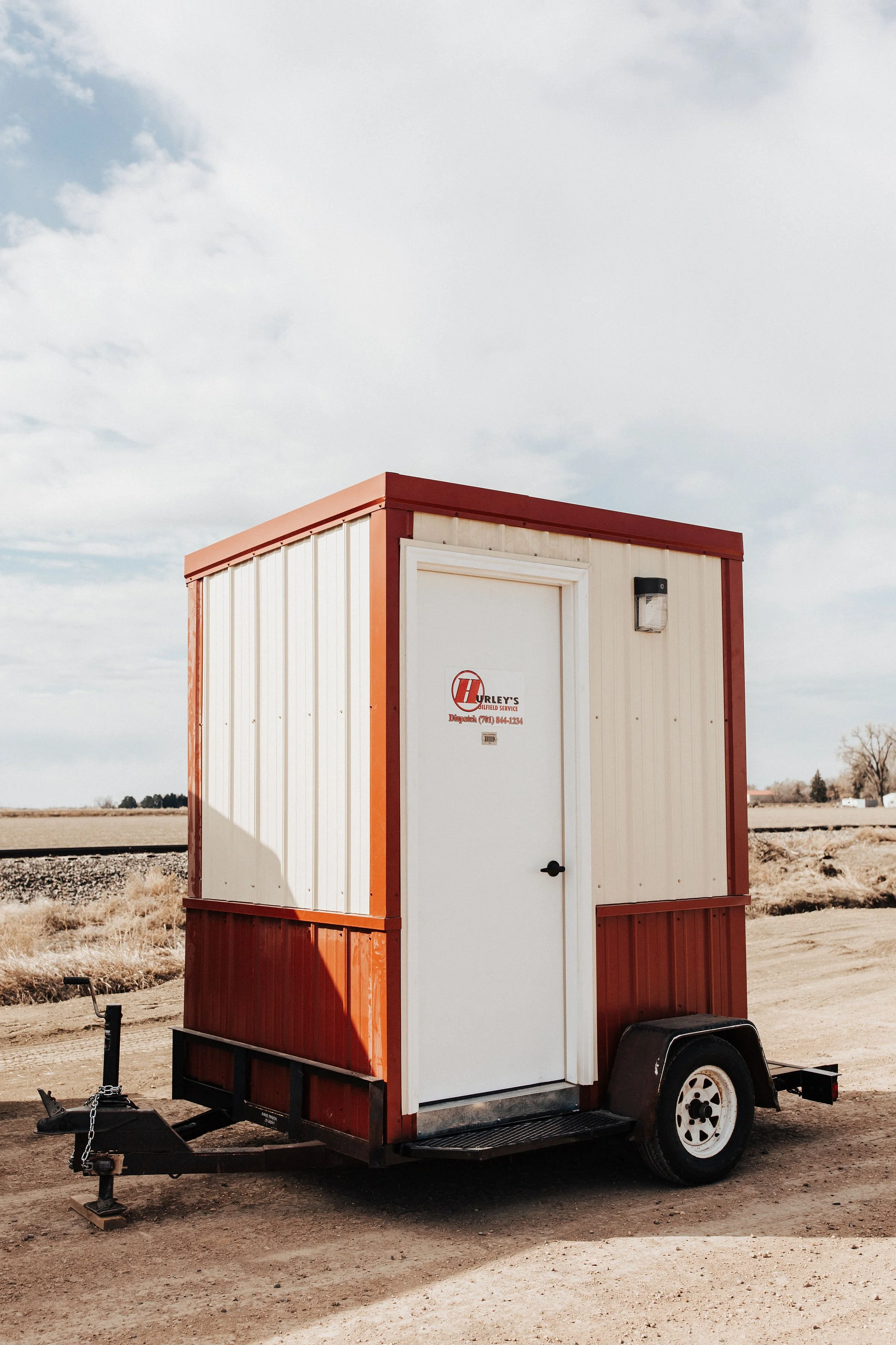 A small mobile service trailer with a white door and red trim, parked on dirt ground under a cloudy sky.