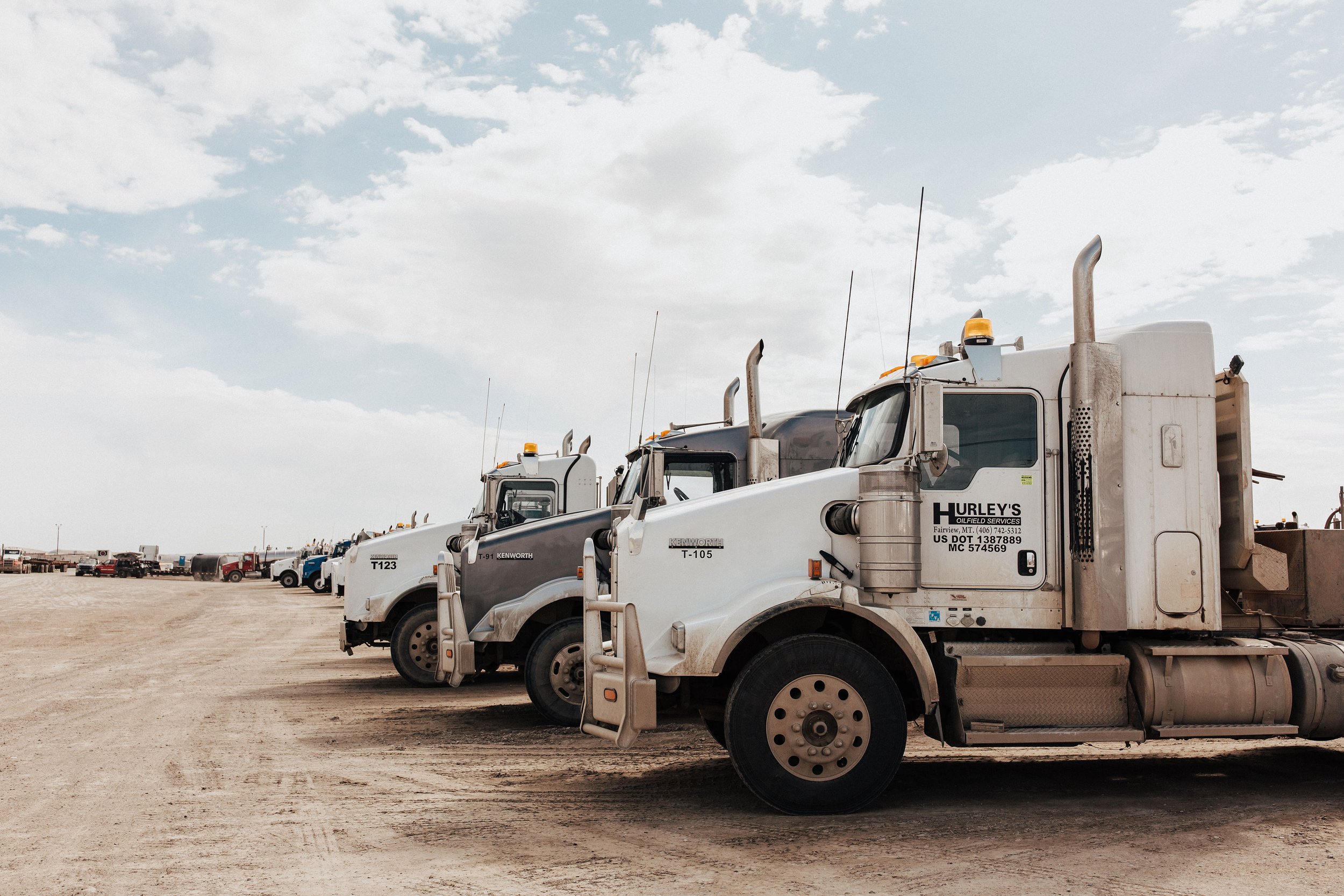 Line of white and gray semi trucks parked on dirt lot under cloudy sky.