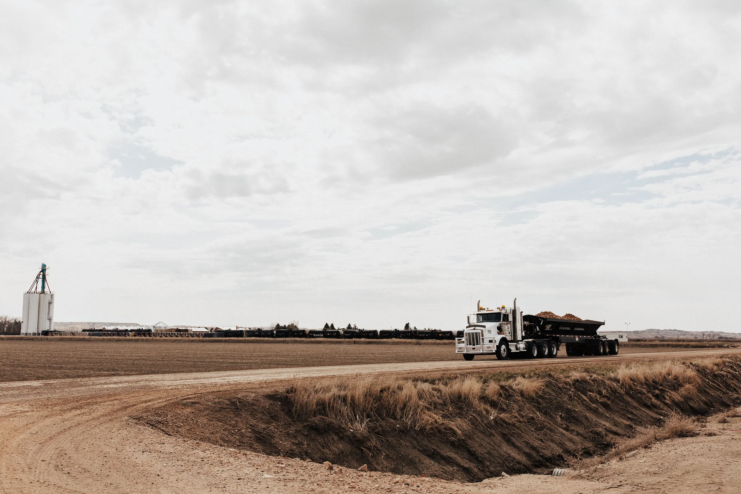 A white semi-truck hauling a black container loaded with dirt driving on a dirt road across an open farmland under a cloudy sky.
