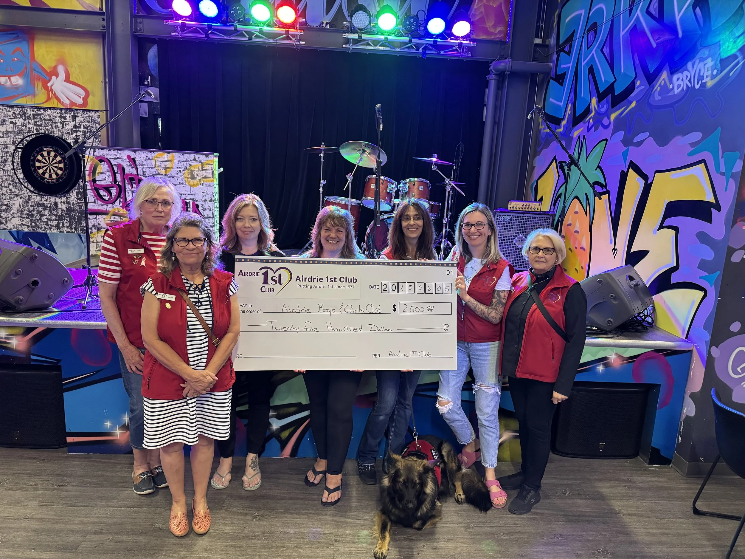 Group of seven women holding a large check on a stage with colorful graffiti background, stage lighting, and musical instruments.