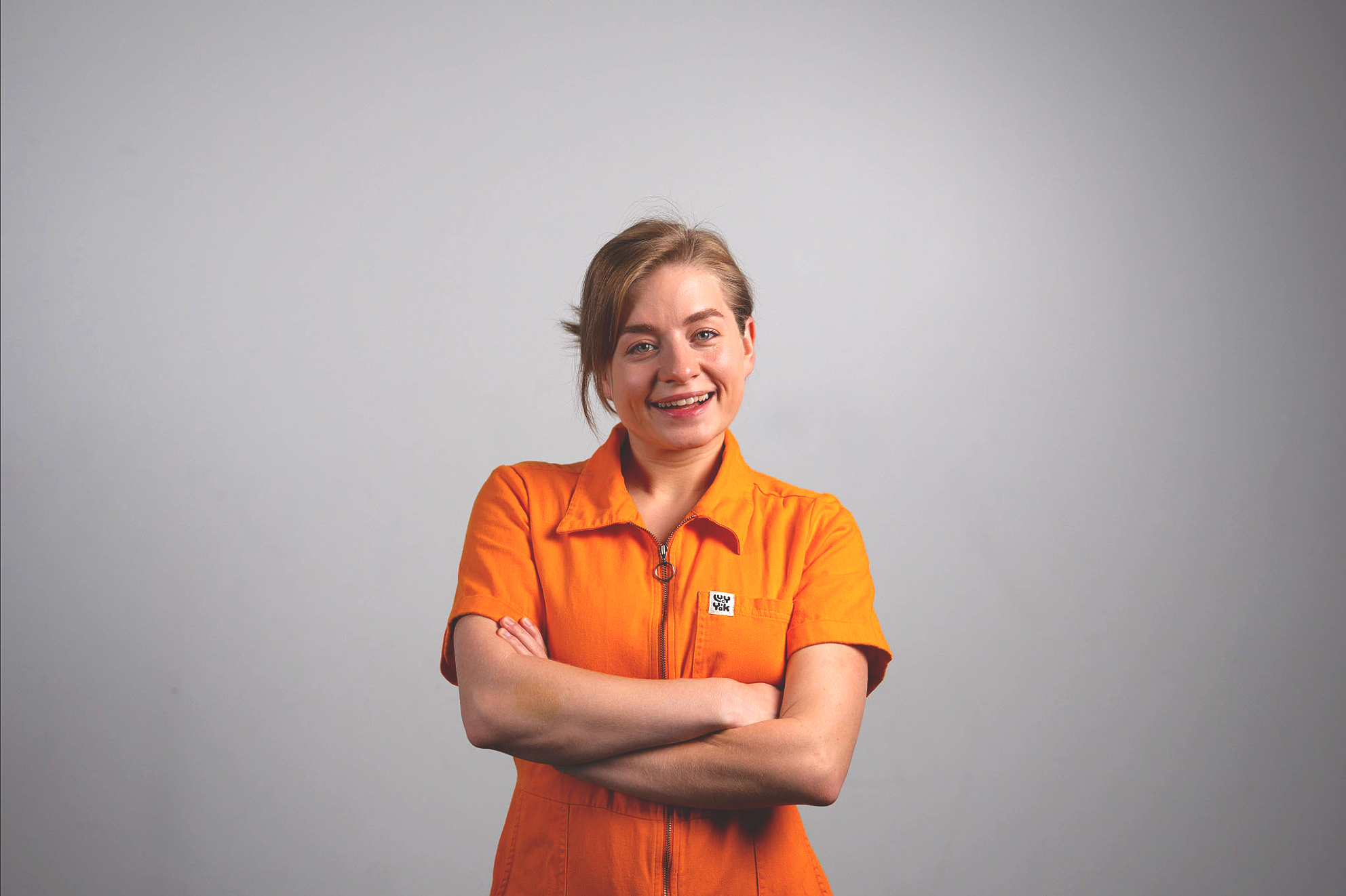 A woman with short brown hair wearing an orange jumpsuit, smiling with arms crossed against a plain gray background.