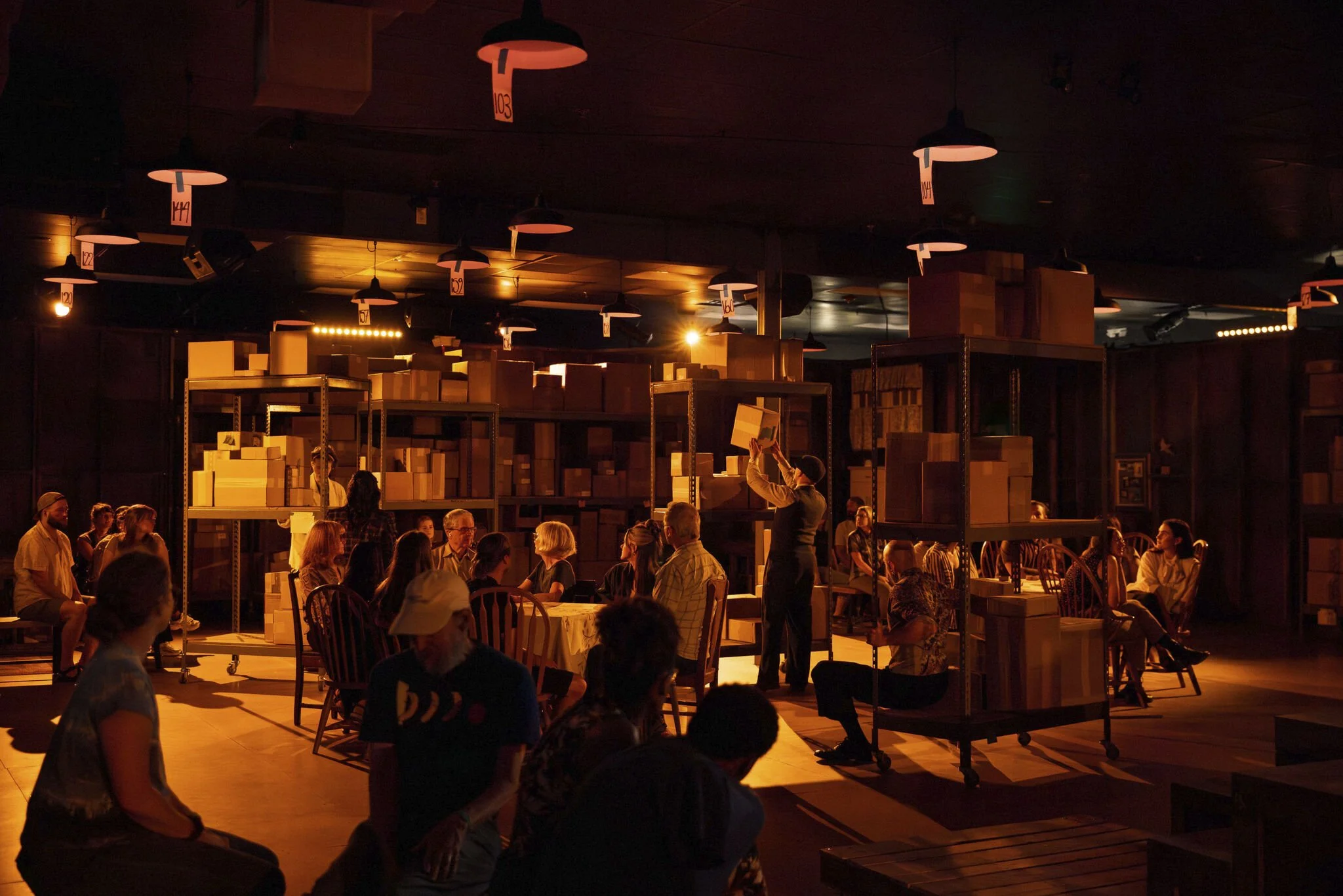 People gathered in a dimly lit venue with warm orange lighting, sitting on chairs at tables and shelves filled with boxes in the background. A person is standing and reaching to pick up a box.